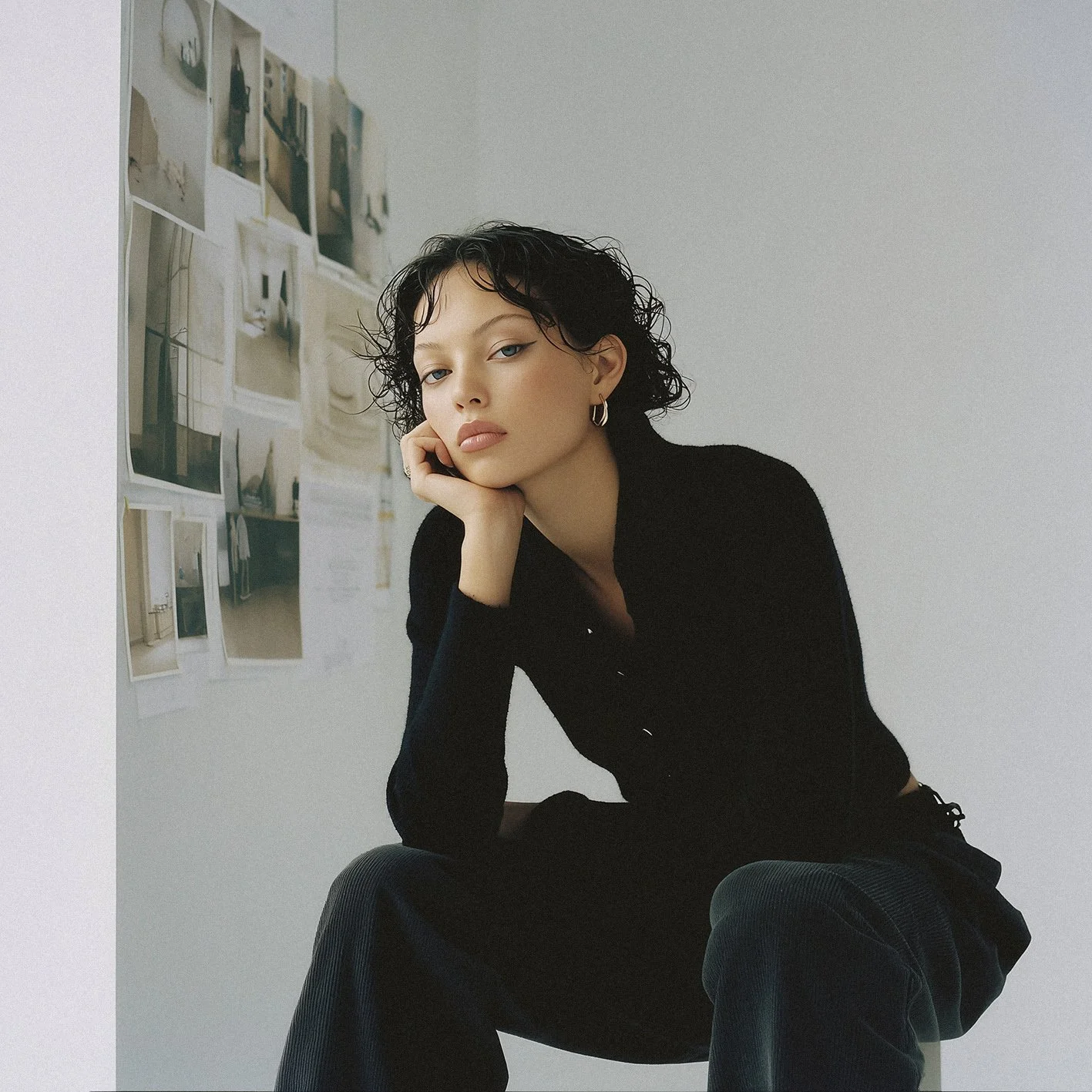Young woman with short curly black hair sitting in front of a white wall with photographs taped on it, wearing a black long-sleeve top and hoop earrings, looking at the camera with a thoughtful expression.