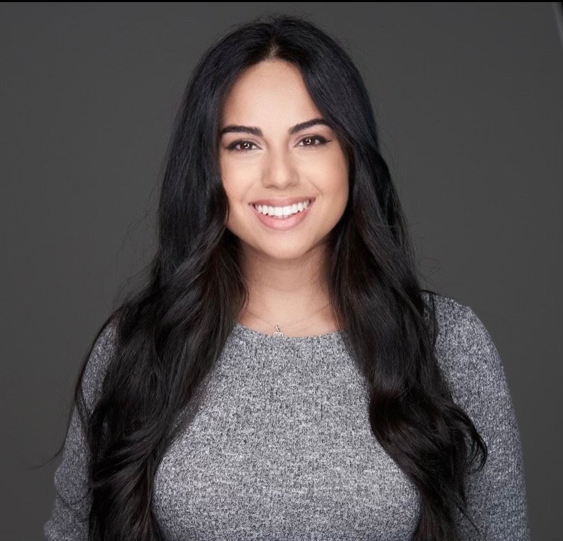 A young woman with long dark hair smiling against a gray background.