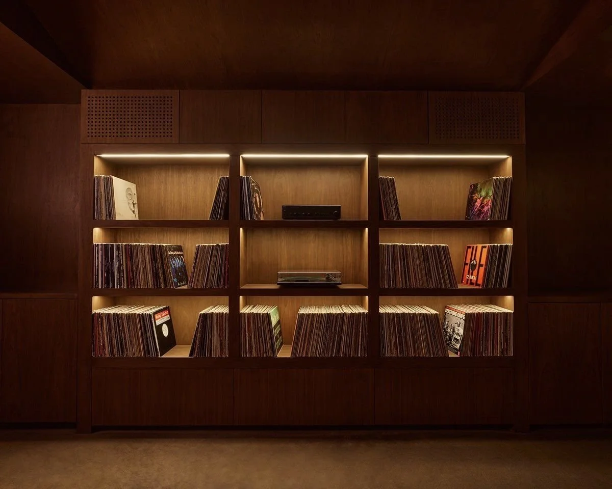 Wooden bookshelf filled with vinyl records, illuminated by internal lighting, set against a dark wood-paneled wall.