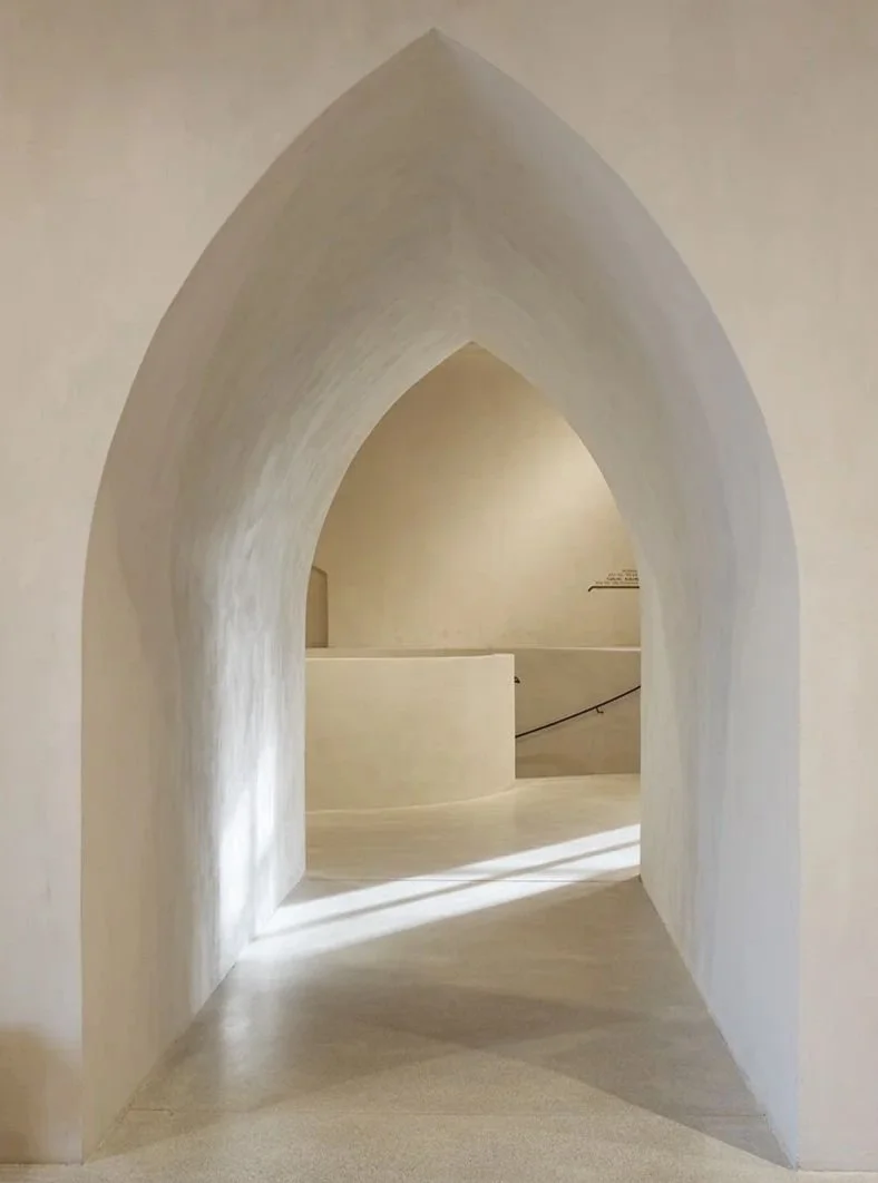 A minimalist, white architectural space featuring a gothic arch doorway leading to a rounded wall and staircase with a white handrail.