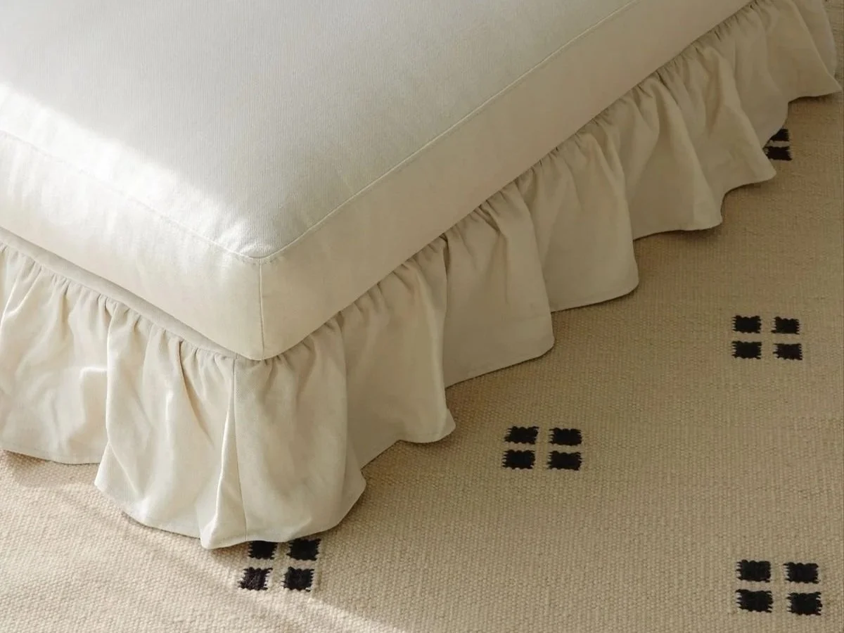Close-up of a cream-colored satin ottoman with gathered skirt on a beige carpet with black square dot pattern.