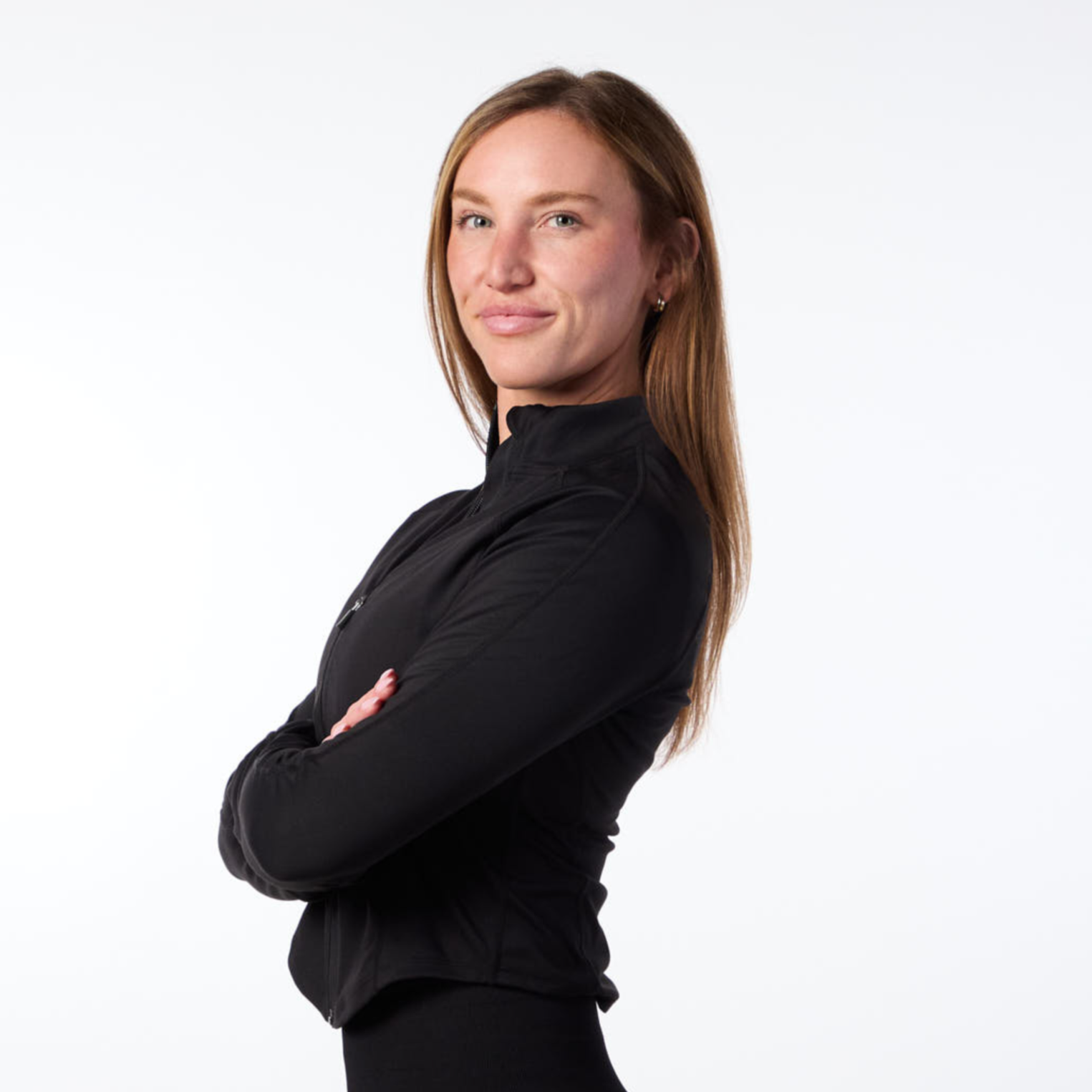 A woman with long reddish-brown hair, wearing a black long-sleeve top, standing with her arms crossed against a plain white background.