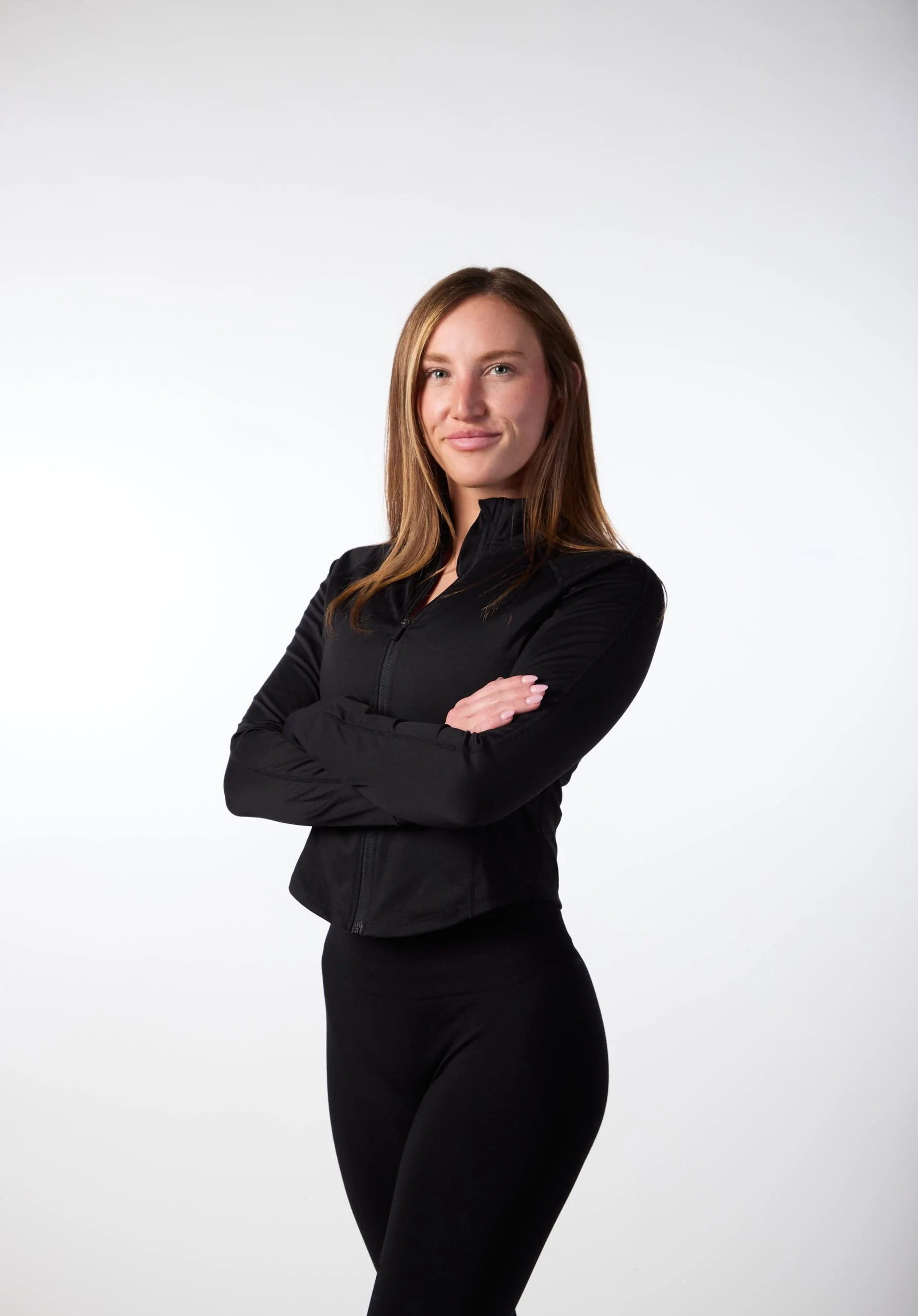 A woman with long brown hair wearing a black athletic jacket and black leggings, standing with arms crossed and smiling in front of a plain white background.