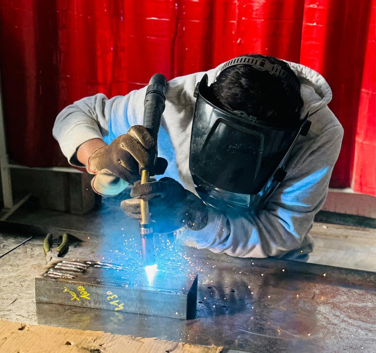 Una persona indossa un grembiule di protezione, guanti e un visore mentre lavora con un saldatore su una piastra di metallo, producendo scintille.