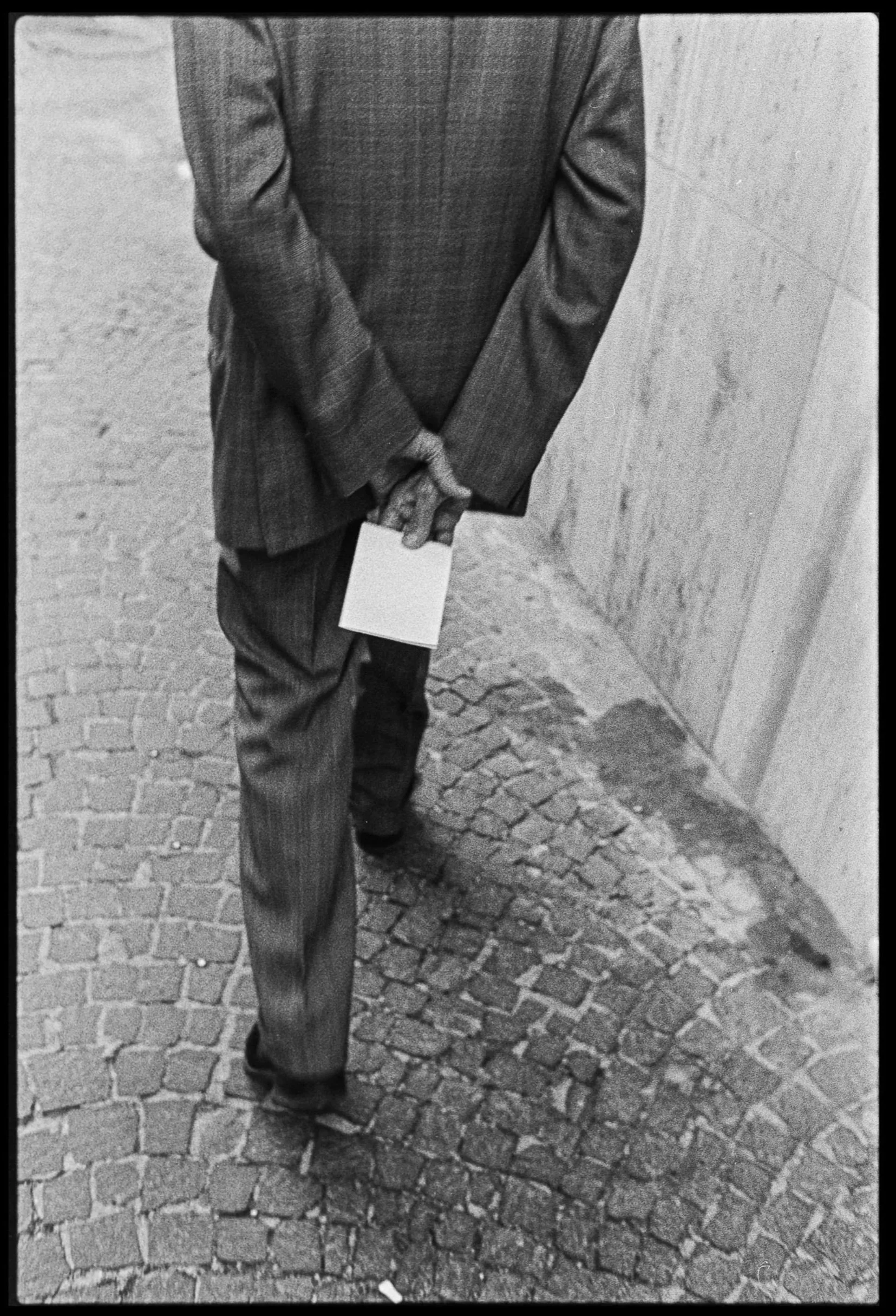 A man in a suit walking on cobblestone pavement, holding a piece of paper behind his back with both hands, near a concrete wall.