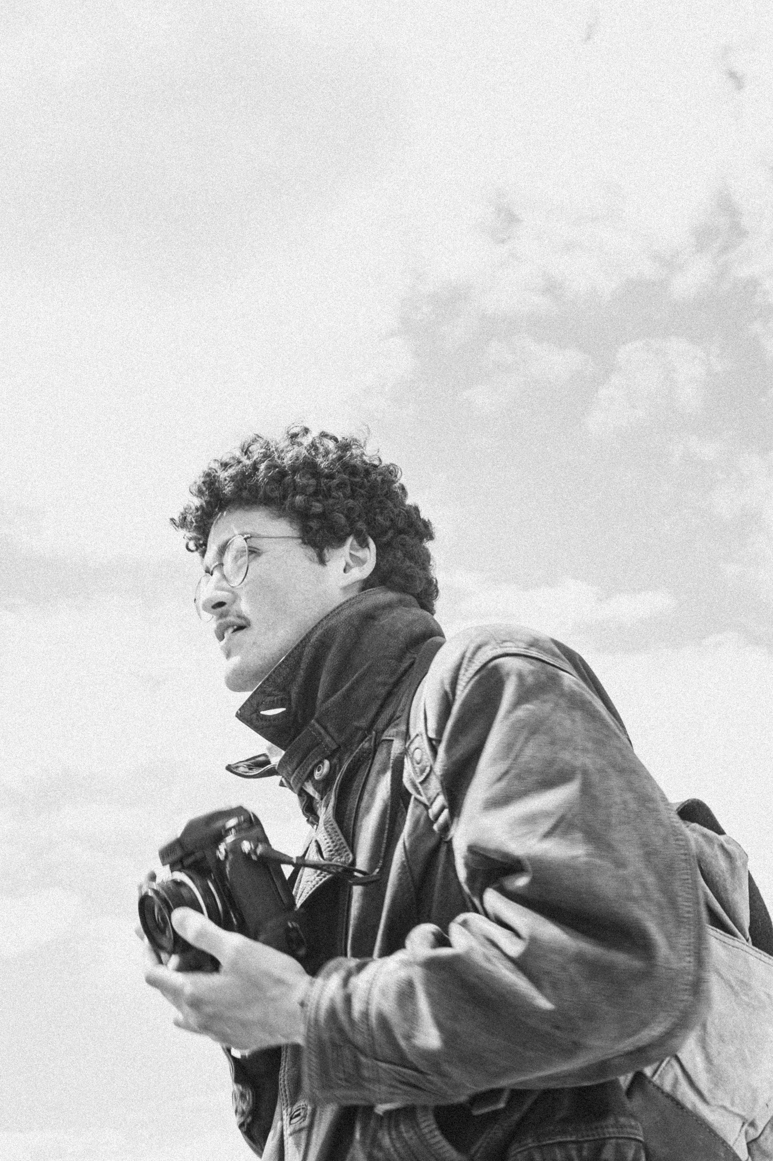 Black and white photo of a man with curly hair and glasses holding a camera, wearing a jacket and backpack, outdoors with cloudy sky.