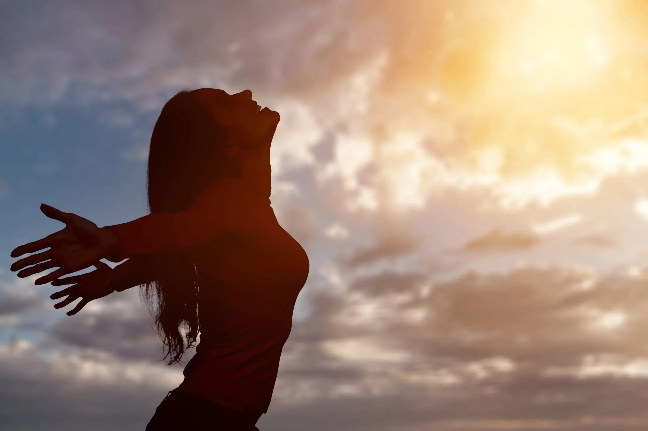 Silhouette of a woman with her arms outstretched, smiling, against a sunset sky with clouds.