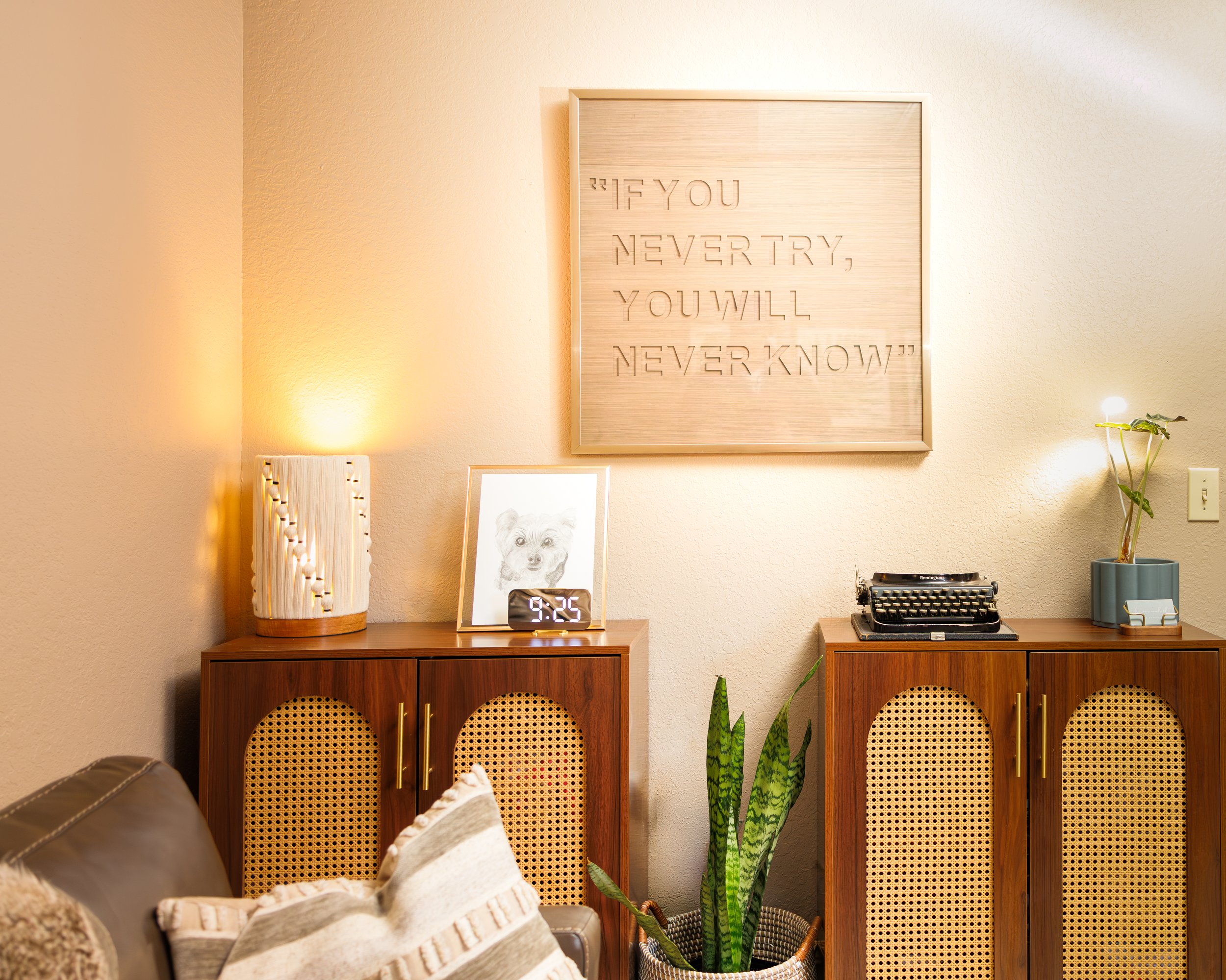 Living room wall with framed artwork and a decorative wooden panel with quote, surrounded by indoor plants, lamps, and furniture.