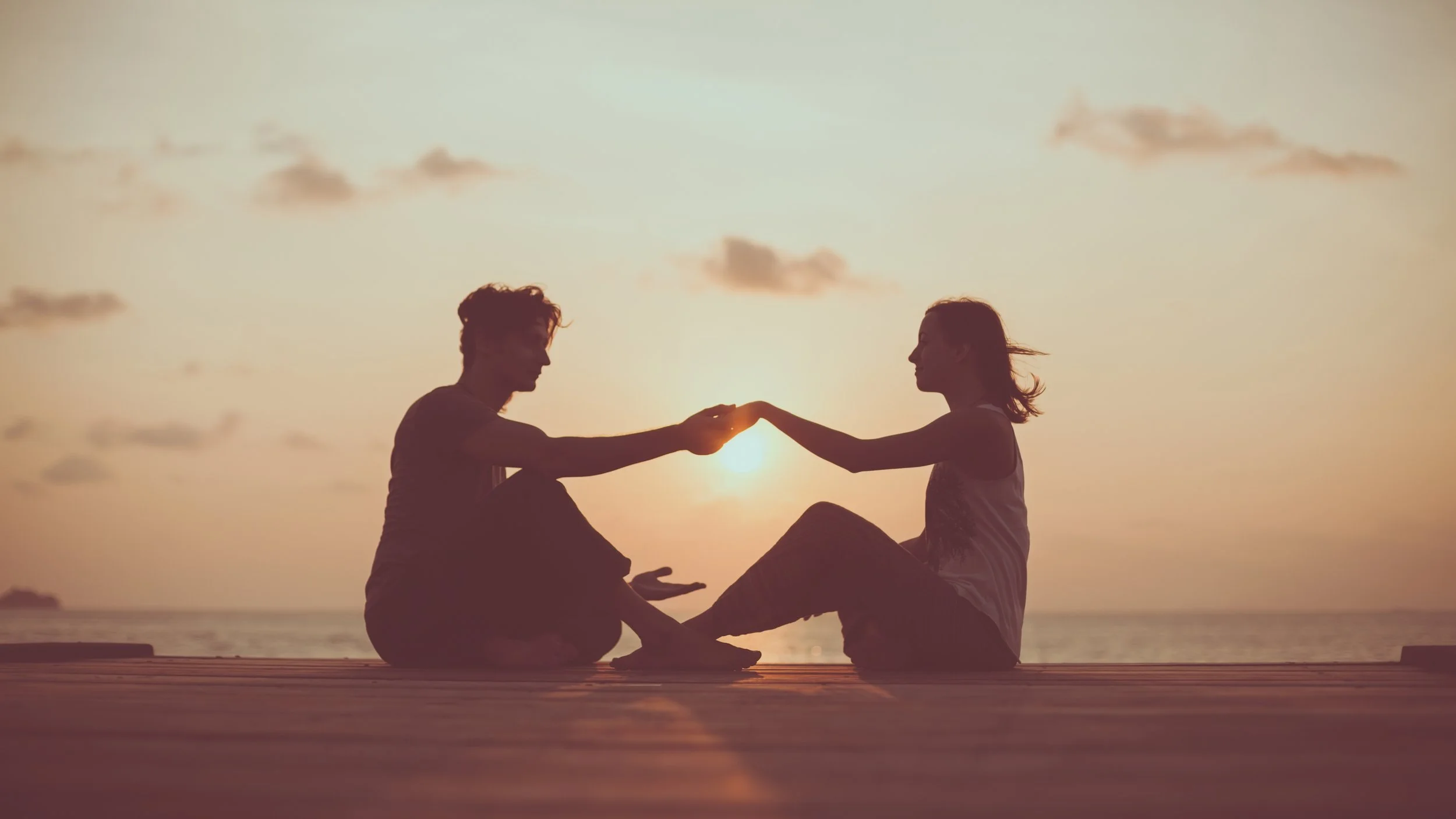 A couple sitting on a deck at sunset, facing each other and holding hands, with the sun setting behind them and a few clouds in the sky.