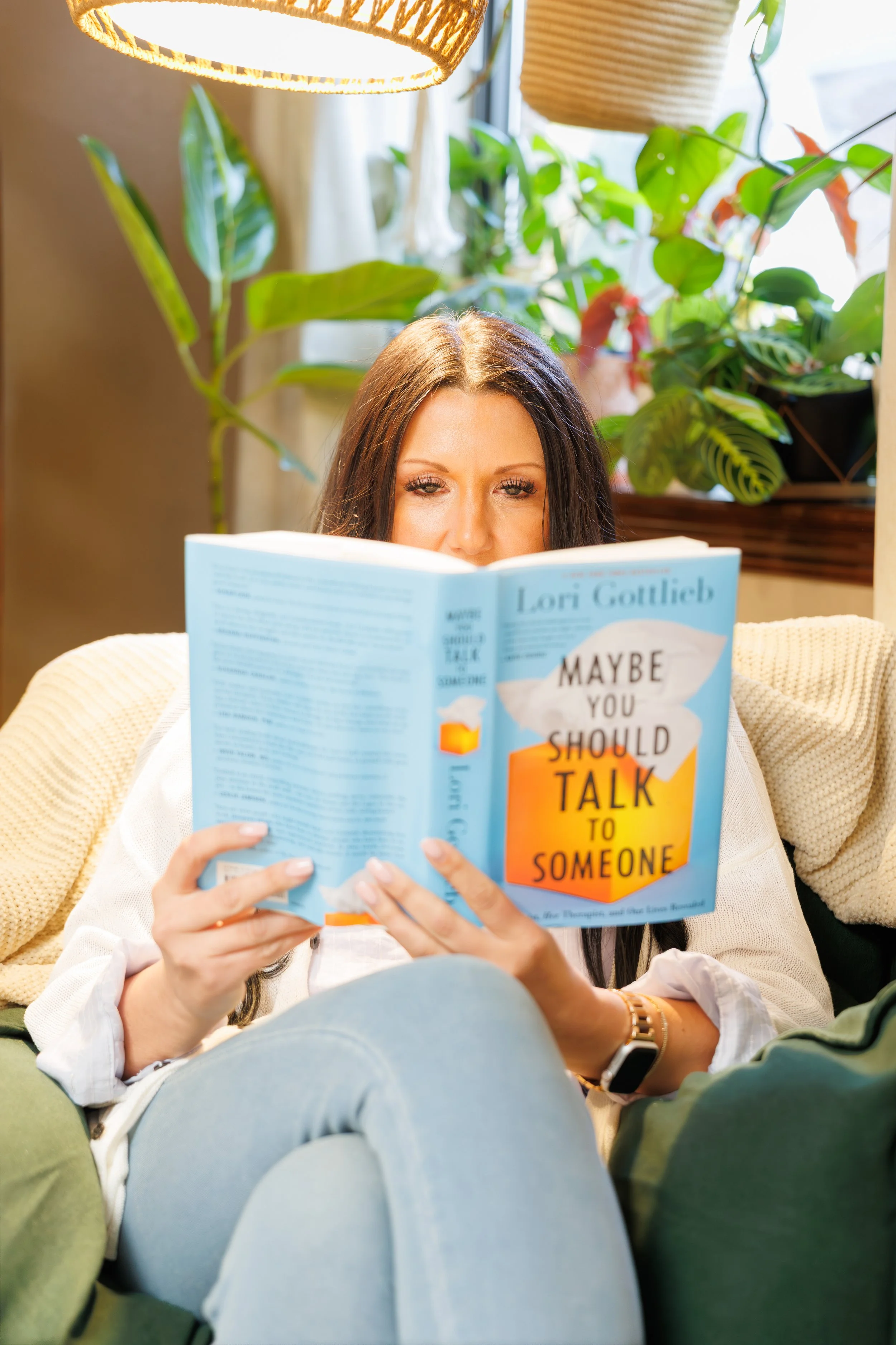 A woman with long dark hair and light skin reading a book titled "Maybe You Should Talk to Someone", sitting on a beige couch with her legs crossed, surrounded by green plants and hanging light fixtures.