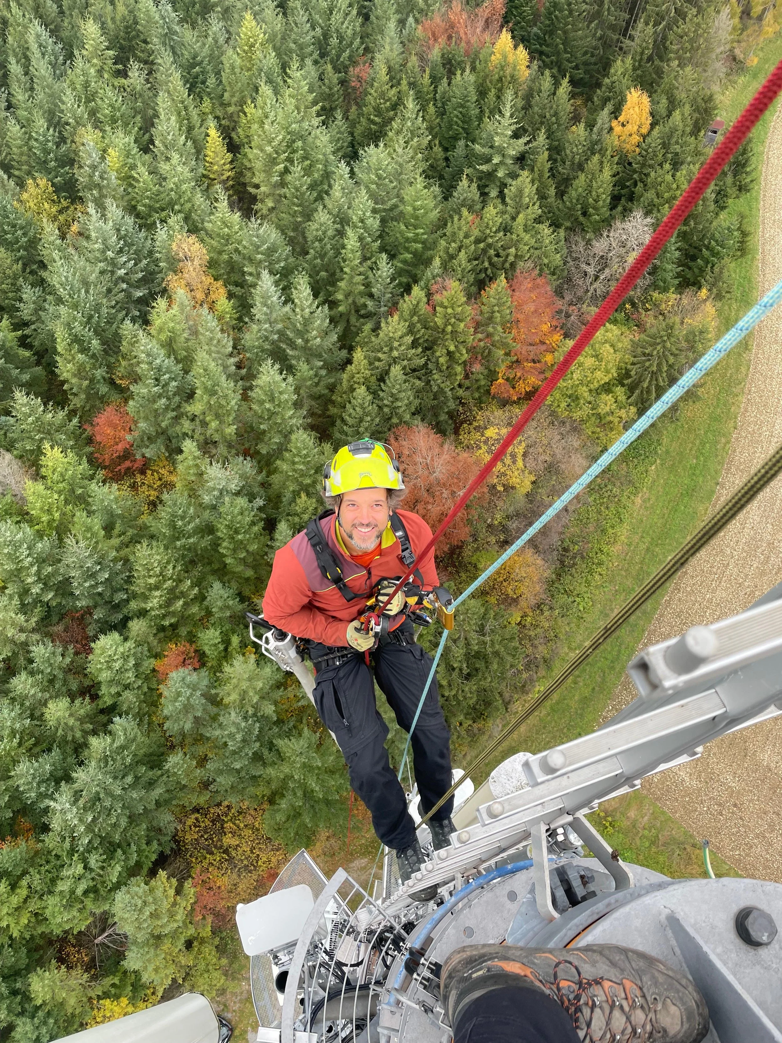 Ein Mann hängt an einem Seil auf einer Leiter hoch oben in einem Baumkronenwald, trägt einen gelben Helm und eine rote Jacke, während er eine Kletterausrüstung benutzt.