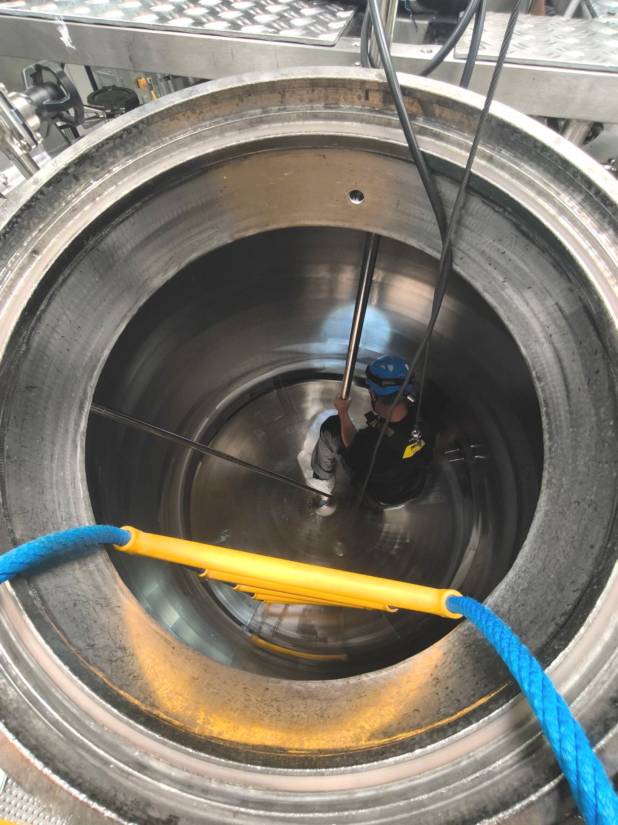 Arbeiter mit Helm und Sicherung in einer großen, metallischen Industrieanlage, möglicherweise einem Rohr oder einer Reaktorhalle, mit Seilen und Sicherheitsvorrichtungen.