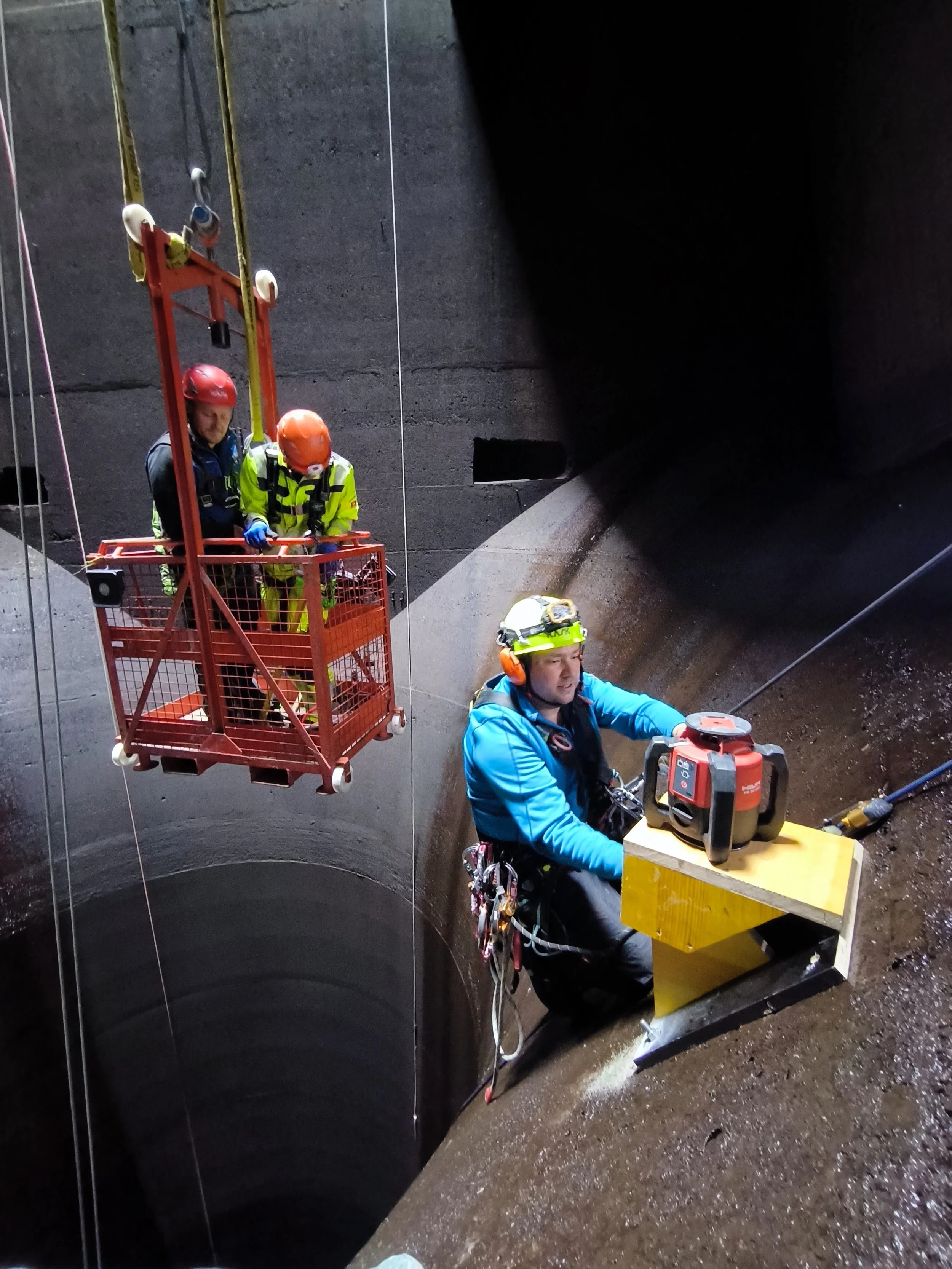 Arbeiter mit Sicherheitsausrüstung arbeitet in einem tiefen, runden Brunnen, während zwei weitere Arbeiter in einem roten Kranwagen auf einer Plattform in der Nähe stehen.