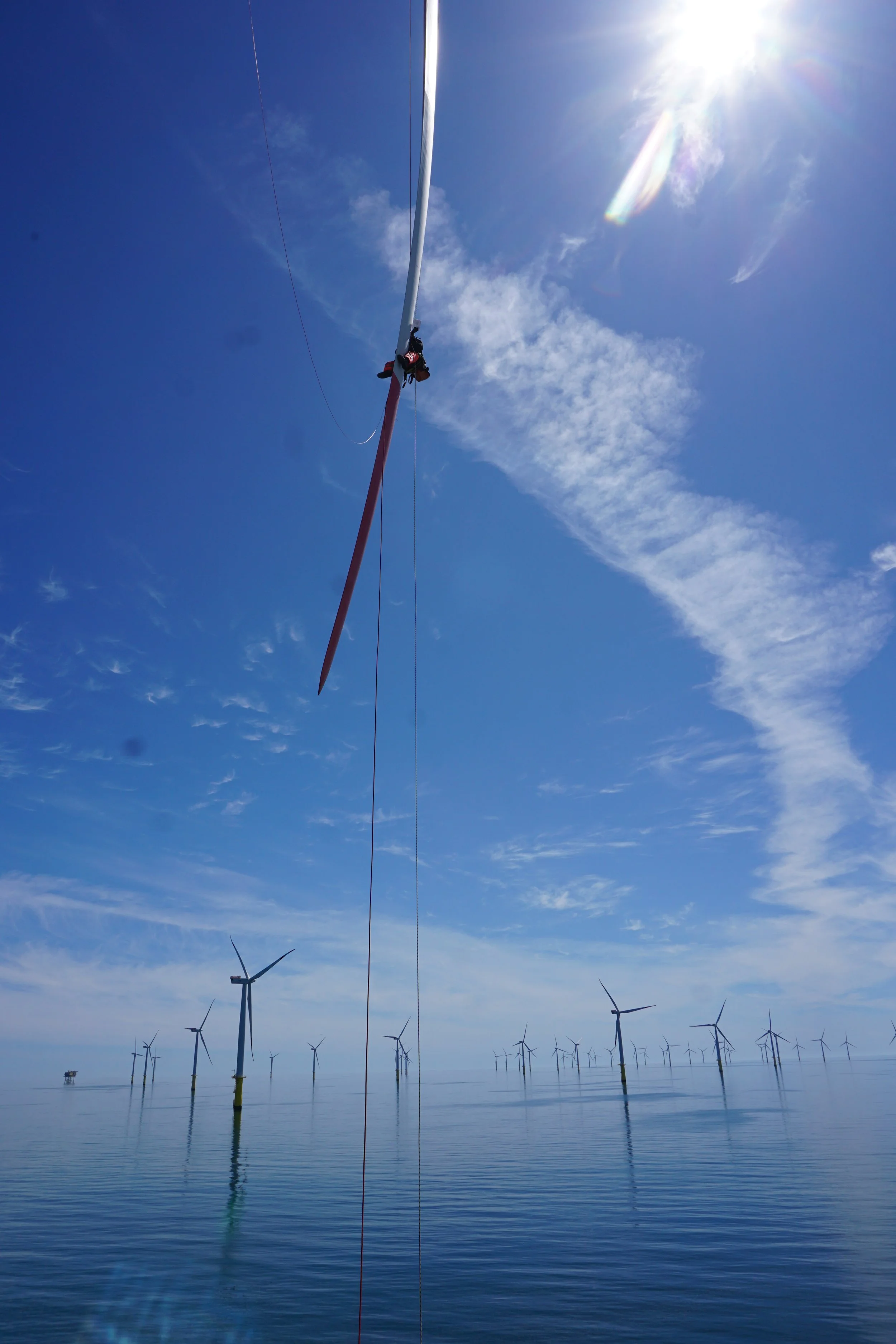Blick auf ein Windpark im Meer mit mehreren Windkraftanlagen, im Vordergrund eine Person, die an einem Windrad arbeitet, unter klarem Himmel mit Sonne