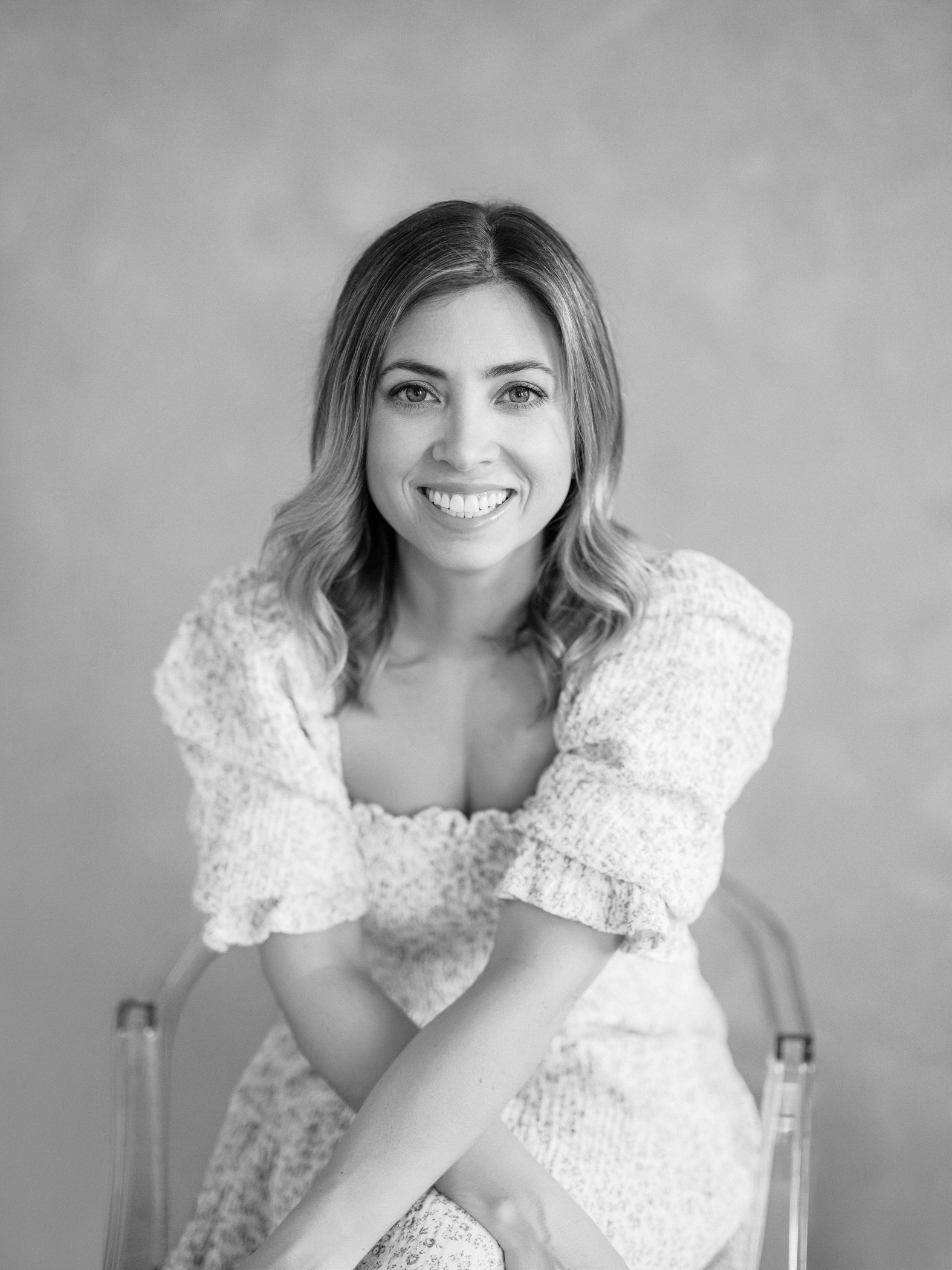 Black and white portrait of a woman smiling, sitting on a chair with arms crossed, wearing a patterned dress, and looking directly at the camera.
