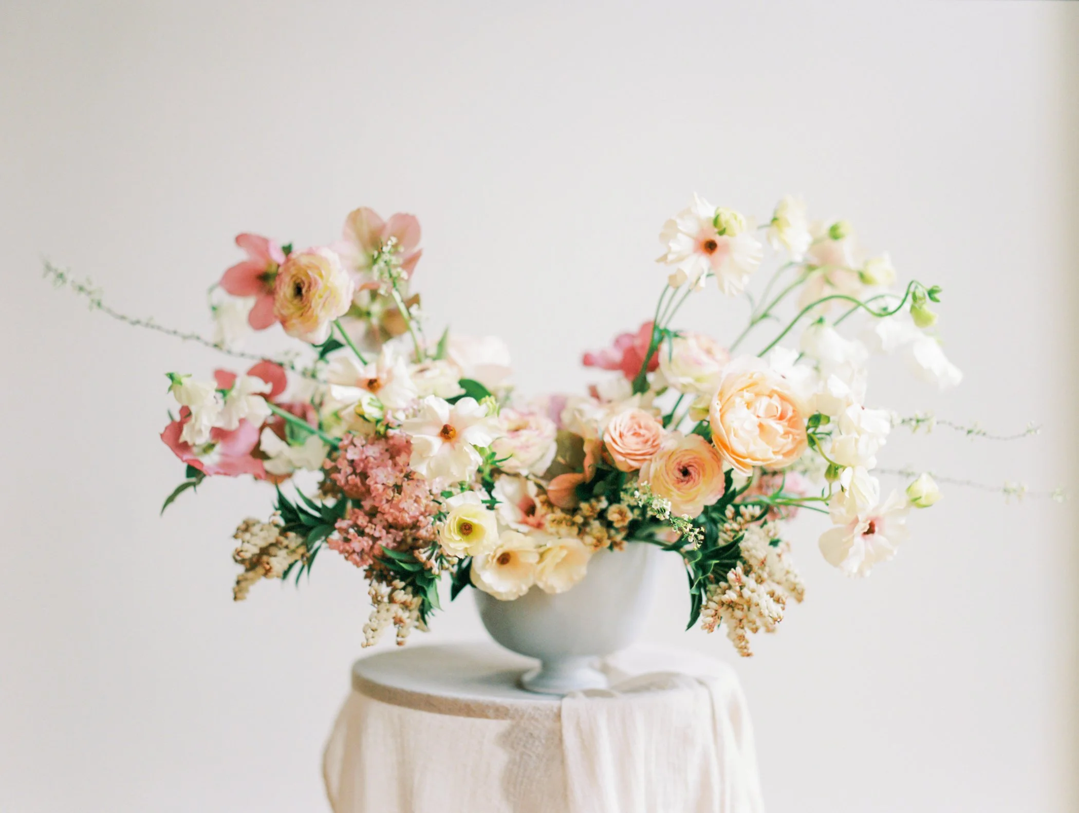 A floral arrangement with pink, peach, and white flowers in a white vase on a wooden pedestal against a plain background.