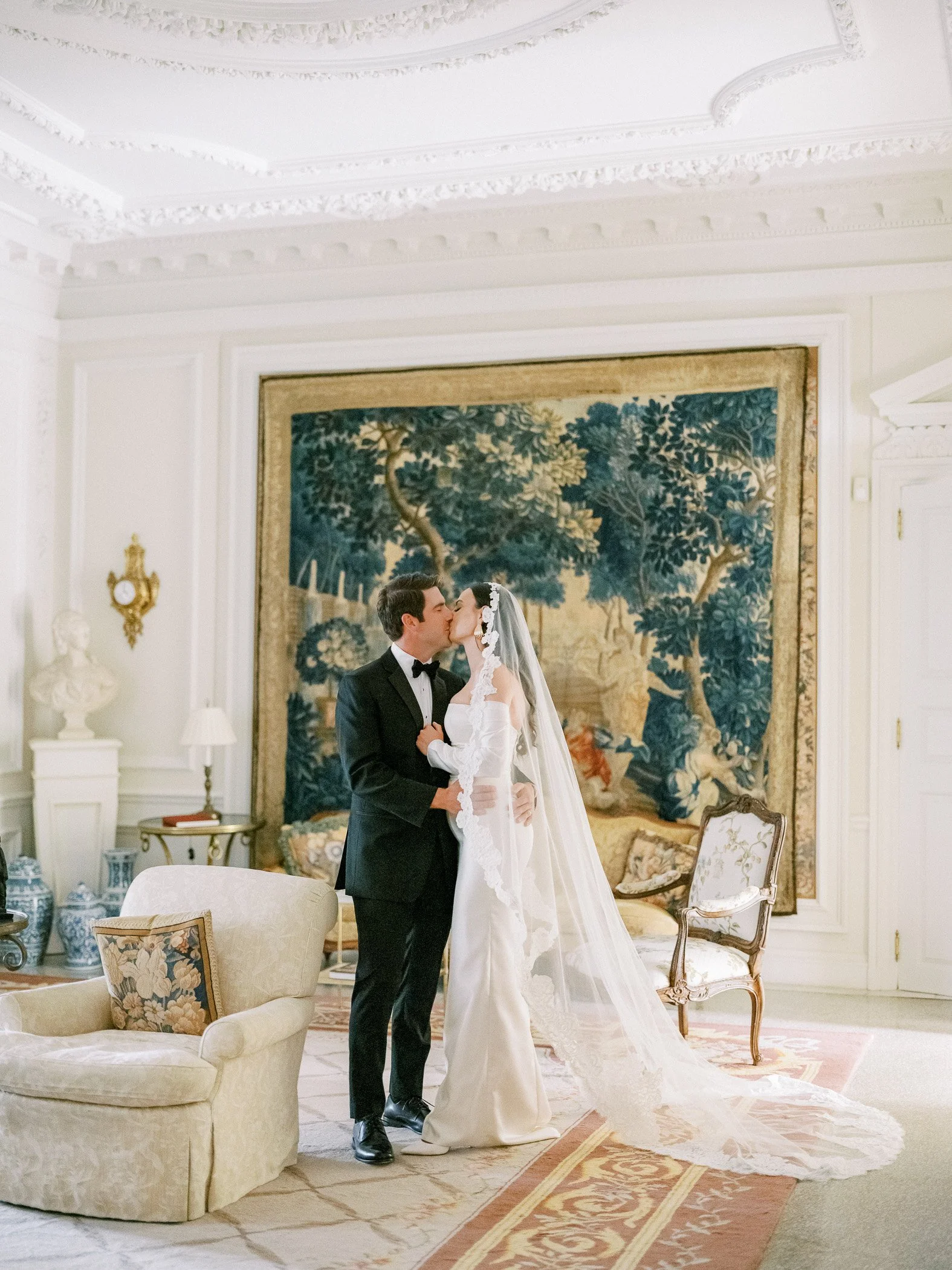 A bride and groom kiss in an elegant room with ornate white walls and a large tapestry behind them. The groom wears a black tuxedo, and the bride wears a white wedding dress with a long veil.