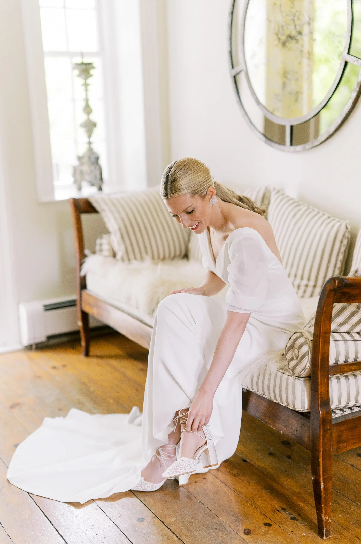 A bride in a white wedding dress putting on white high-heeled shoes with lace details.