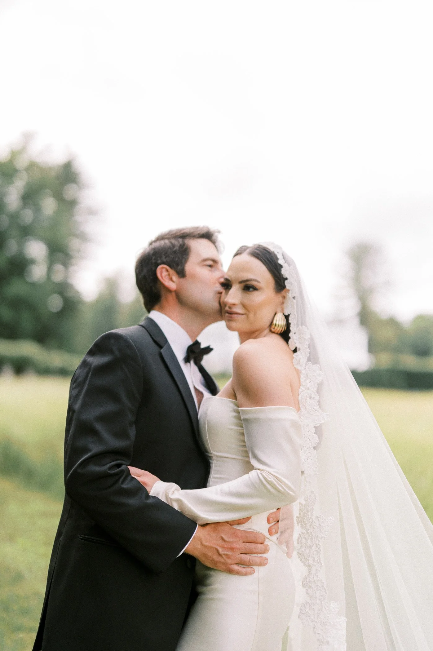 A newlywed couple in wedding attire outdoors; the groom in a black tuxedo kisses the bride's temple, who wears an off-shoulder wedding gown with lace details and a veil.