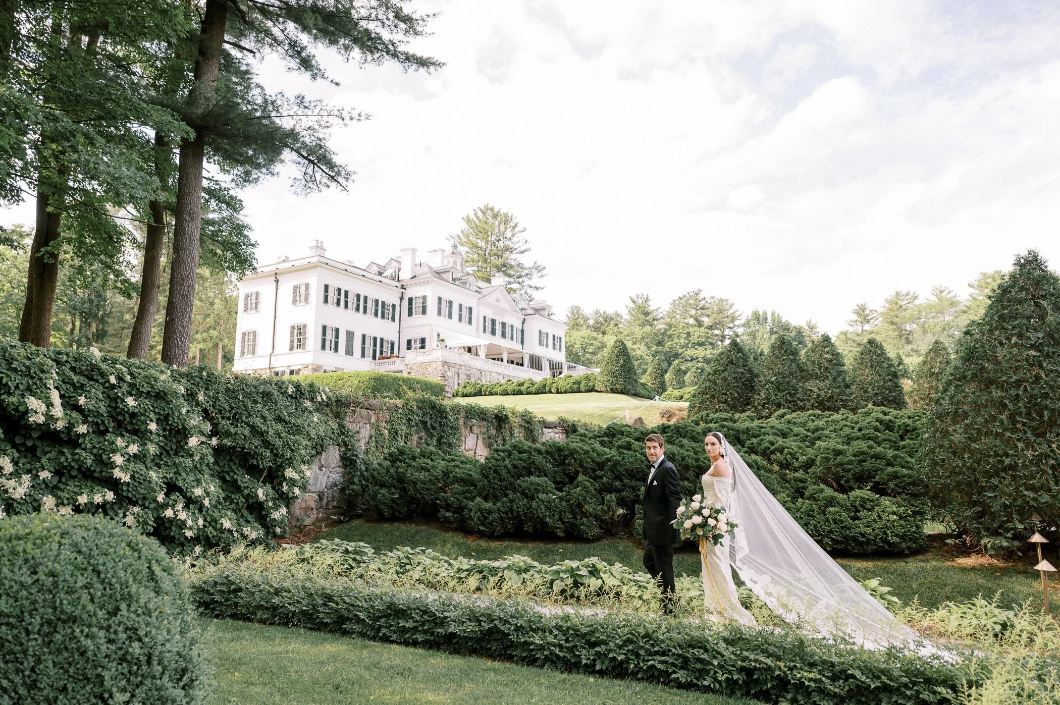 A bride and groom stand outdoors in a lush garden with green bushes and trees, holding a bouquet of flowers, with a large white mansion in the background.