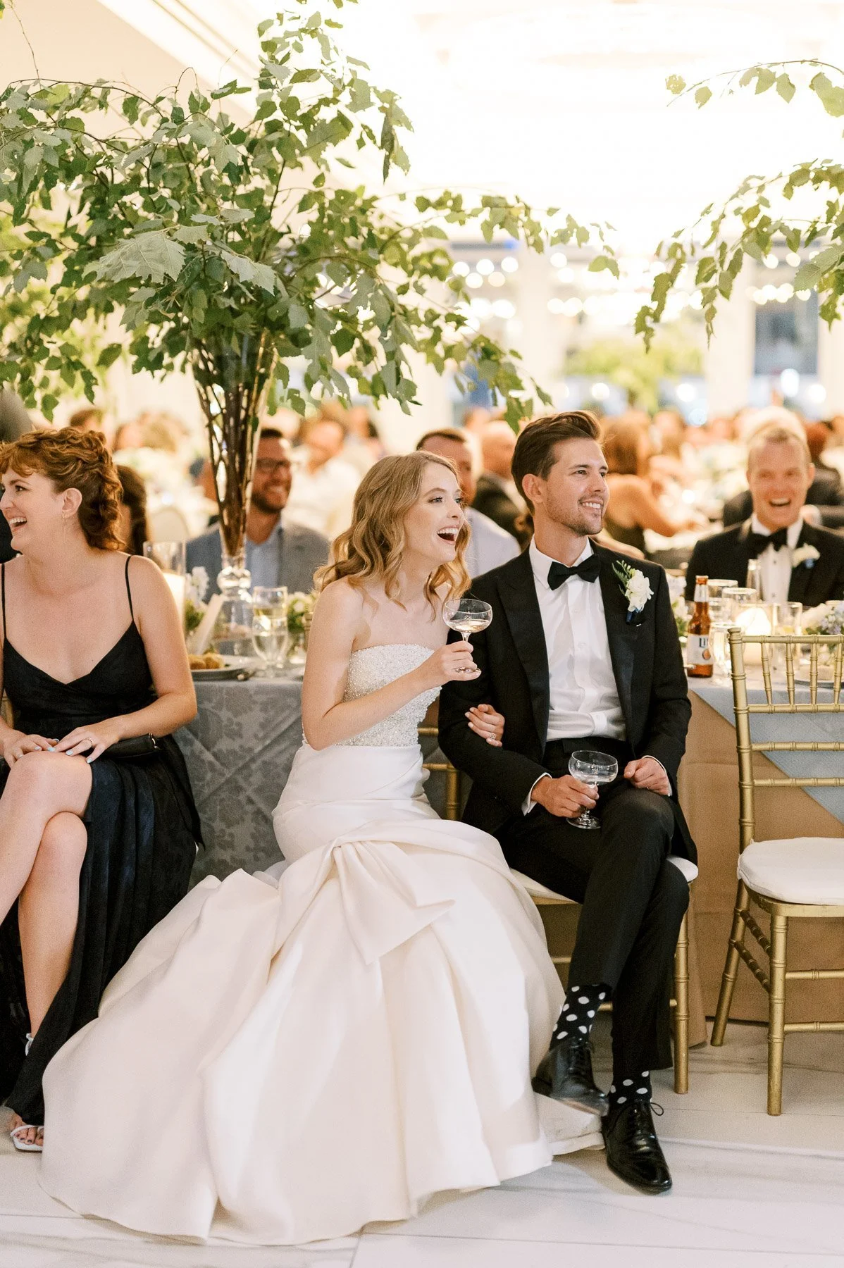 A bride and groom at a wedding reception sitting together, smiling, with guests in the background. The bride wears a white strapless wedding gown, and the groom wears a black tuxedo with a bow tie and boutonniere. They are seated at a decorated table