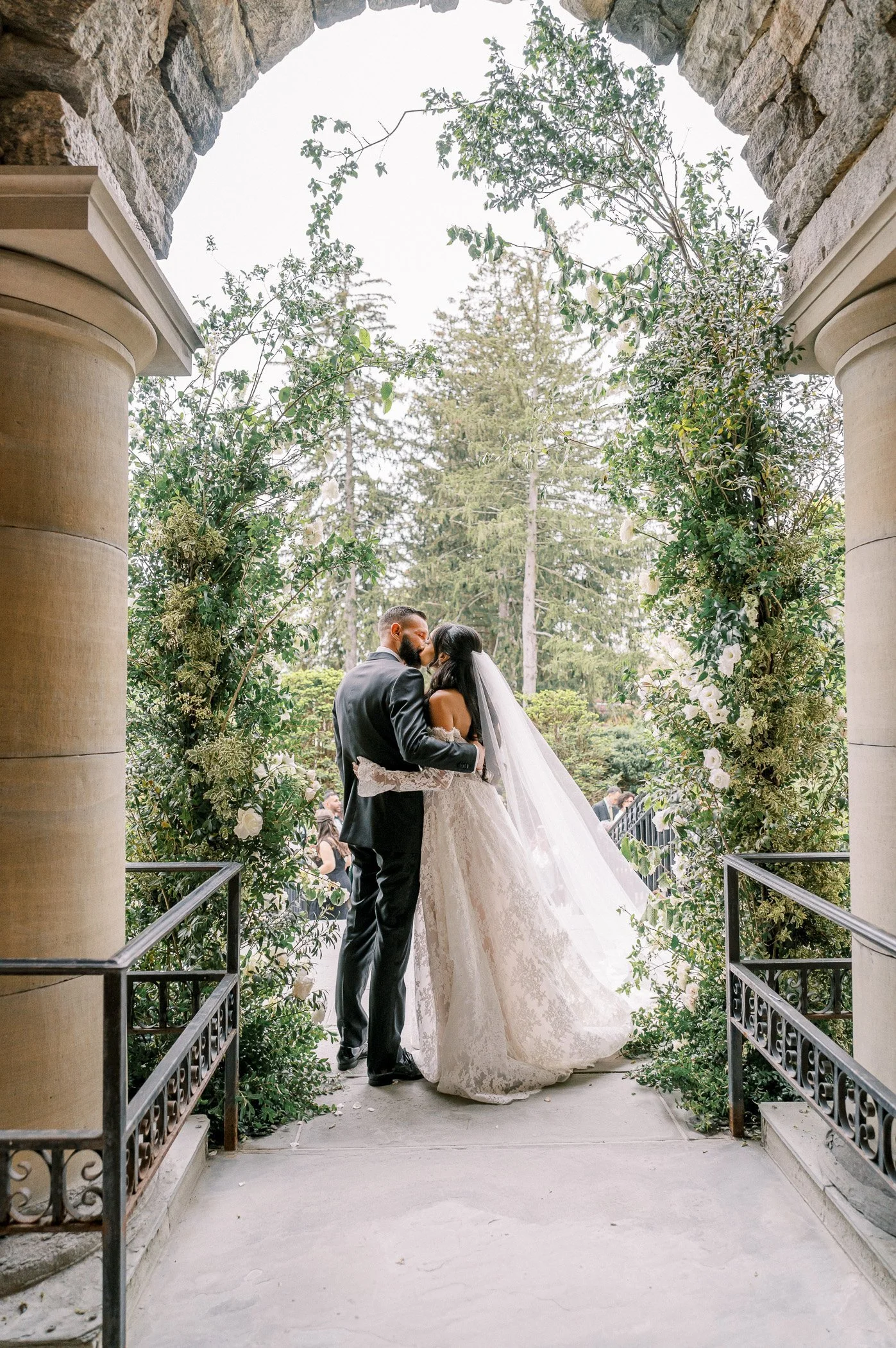 A bride and groom sharing a kiss during their wedding ceremony beneath a stone archway decorated with green foliage and white flowers.