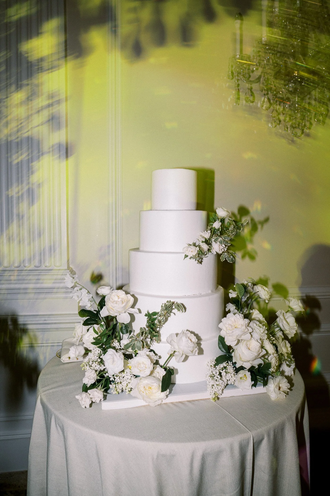 A four-tiered white wedding cake decorated with white flowers and green foliage, displayed on a round table covered with a cream-colored tablecloth.