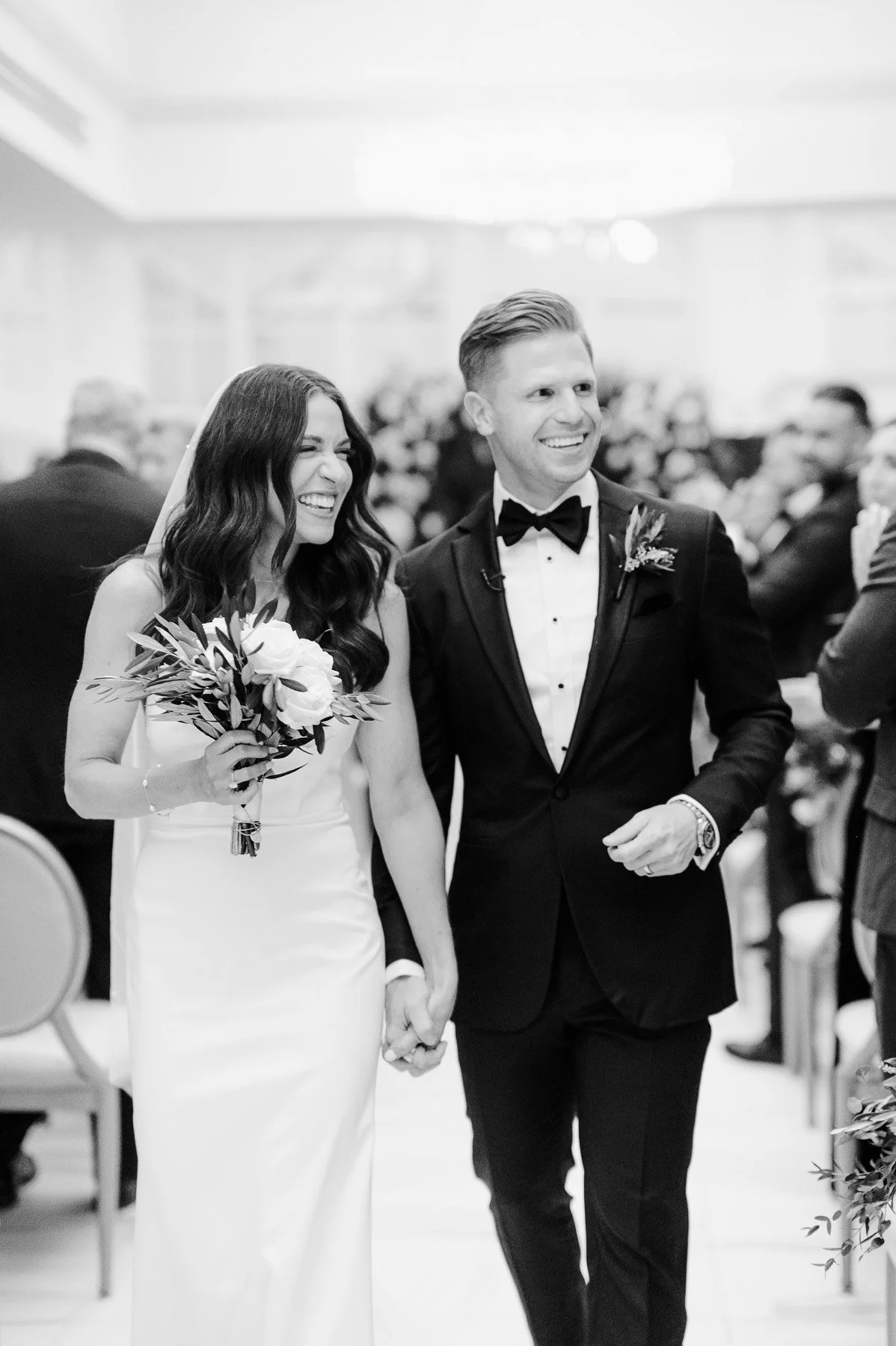 Black and white photograph of a bride and groom walking down the aisle during their wedding ceremony, with the bride smiling and holding a bouquet, and the groom smiling and looking forward, surrounded by guests