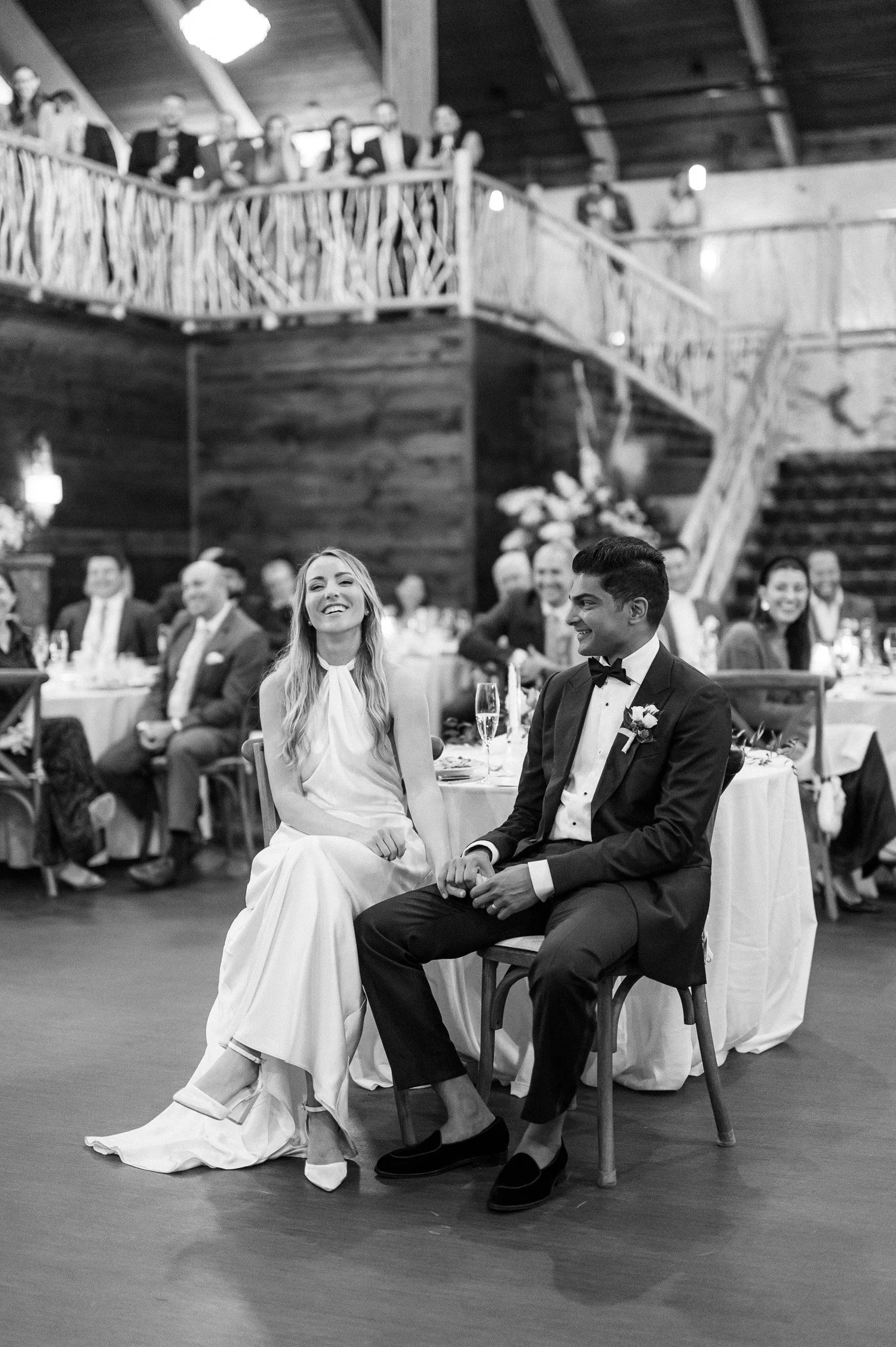 A black and white photo of a bride and groom sitting closely at their wedding reception, holding hands, with guests seated at tables in the background.