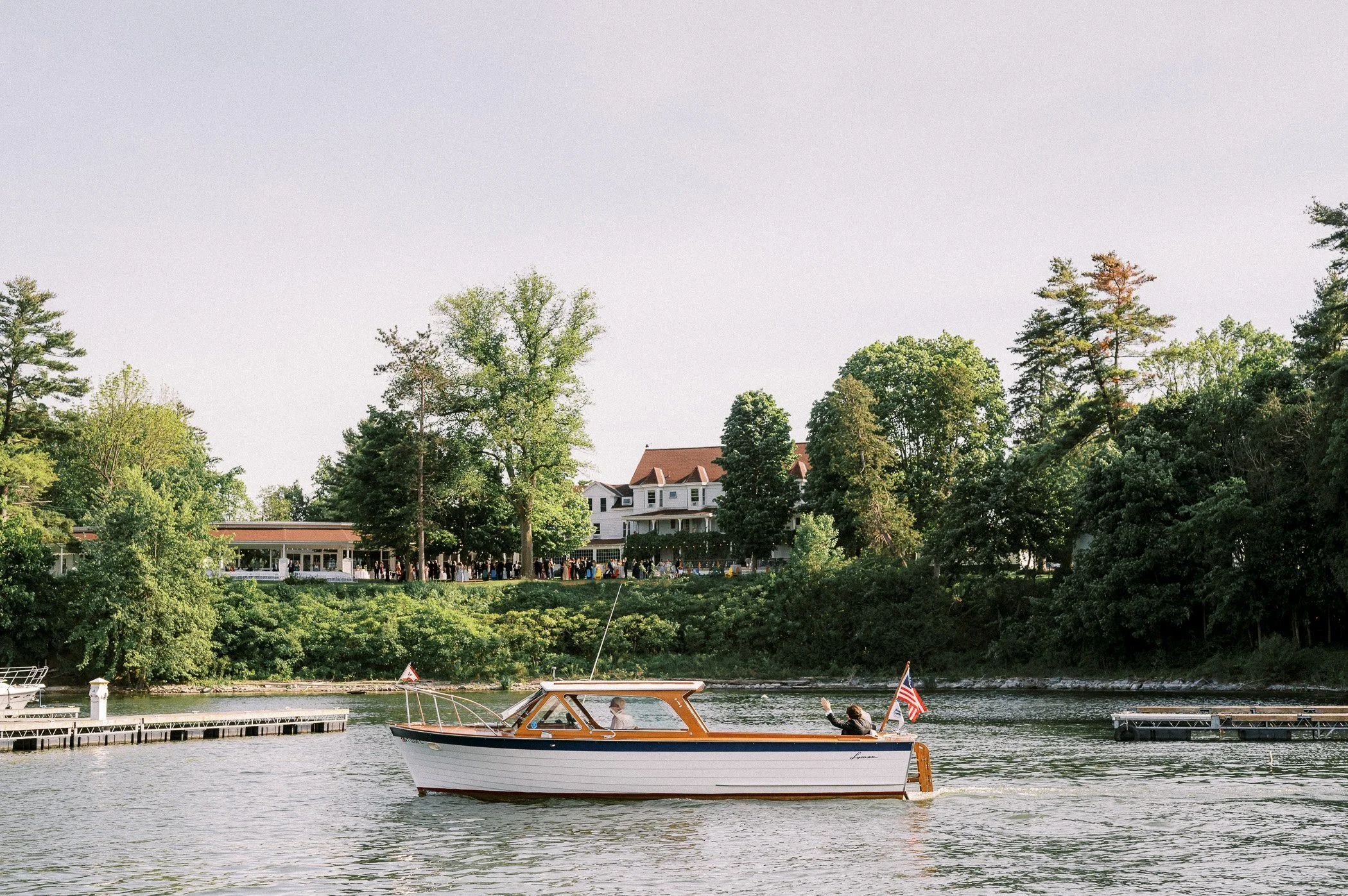 A boat with two people on a river, surrounded by green trees and houses on the riverbank.
