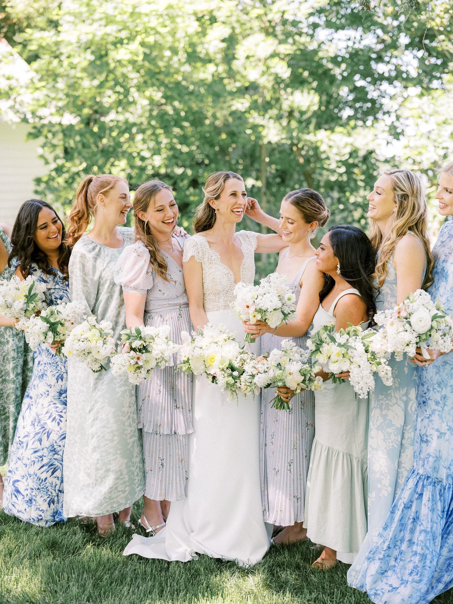 A bride surrounded by bridesmaids during an outdoor wedding on a sunny day, all holding bouquets of white flowers and smiling.