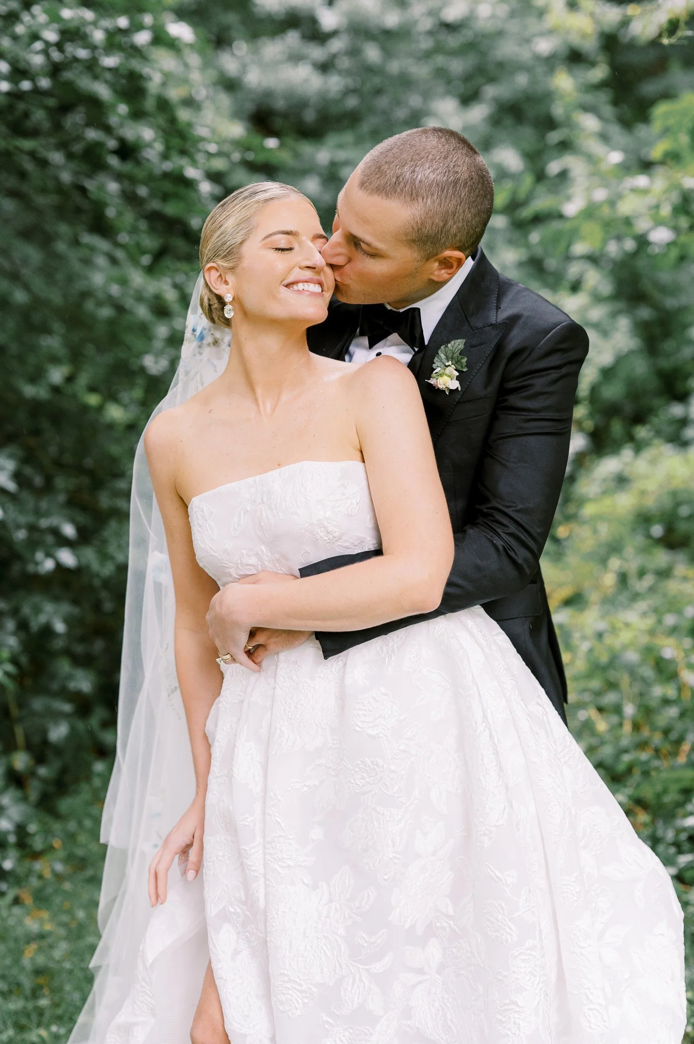 A newlywed couple sharing a romantic kiss outdoors, with the groom in a black tuxedo and the bride in a white wedding dress, surrounded by greenery.