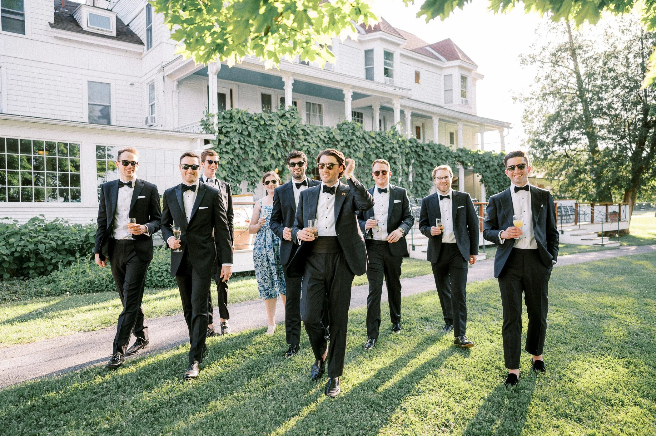 A group of men in tuxedos and women in dress walking on a lawn outside a large white house, holding drinks and smiling in the sunlight.