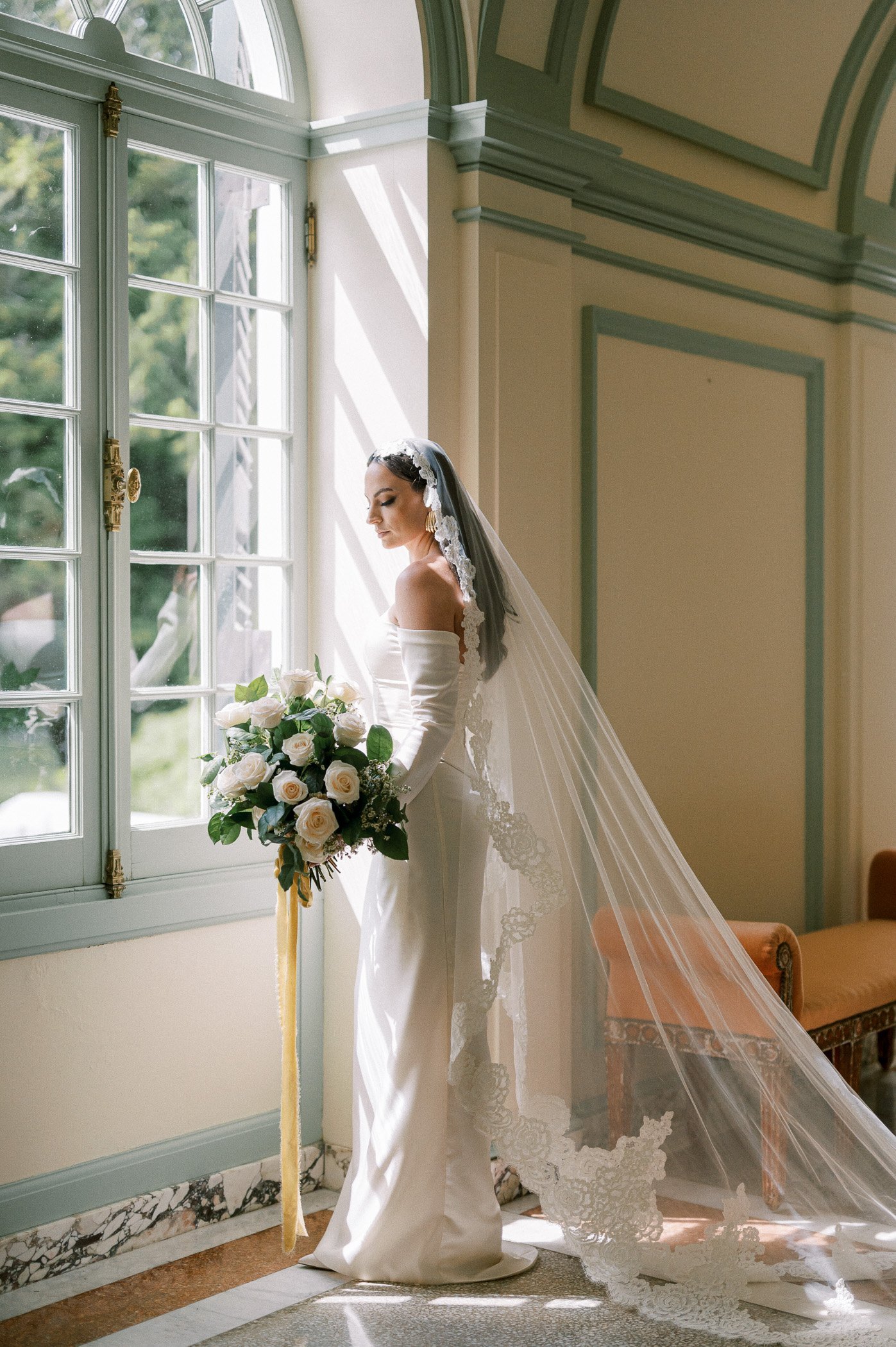 A bride in a white off-shoulder wedding gown and veil stands by a window, holding a bouquet of white roses with green foliage, inside a decorated room with pastel-colored walls and a marble floor.