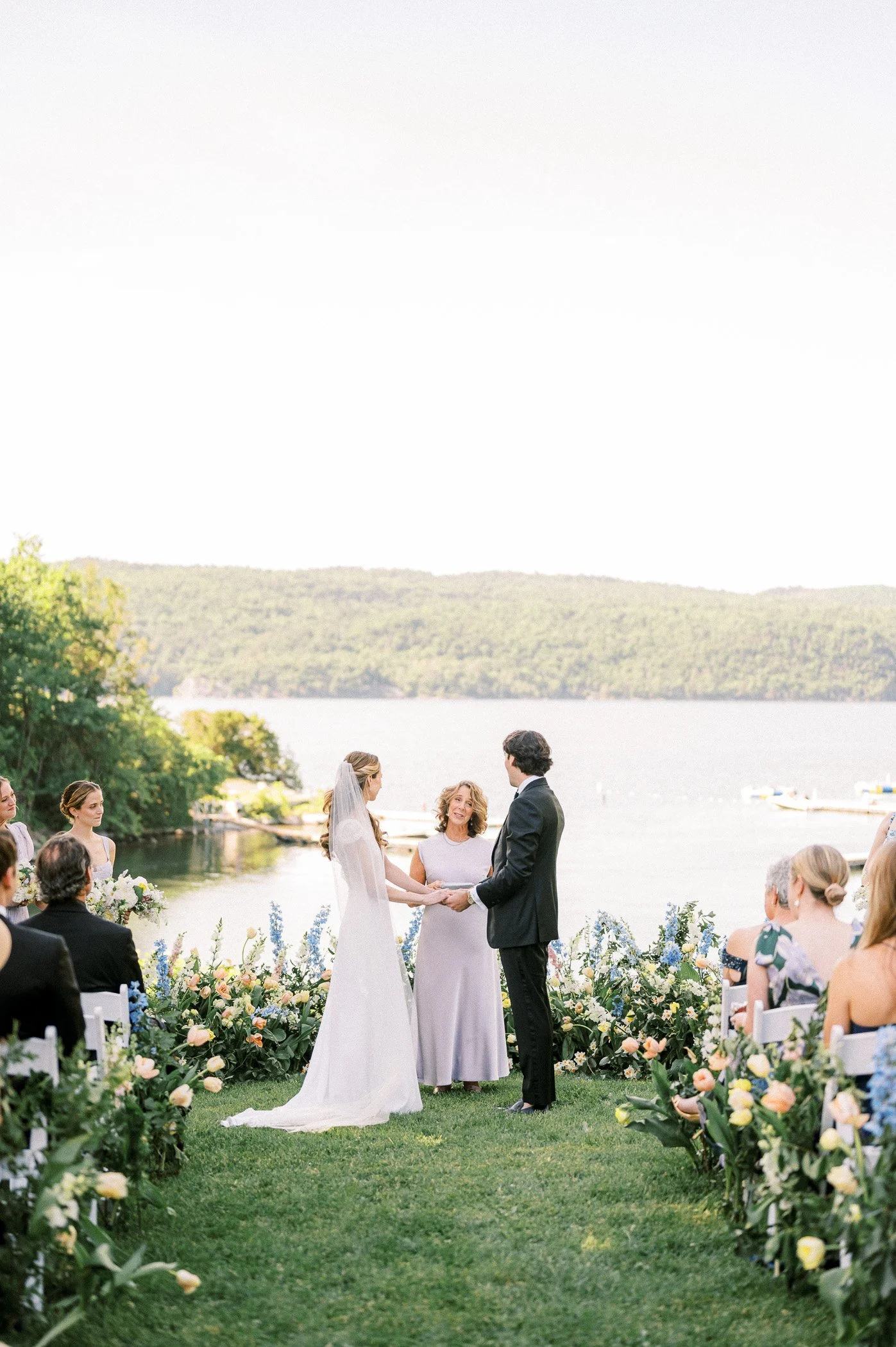 A wedding ceremony outdoors by a lake with a bride and groom holding hands, surrounded by friends and family, with scenic green hills in the background.