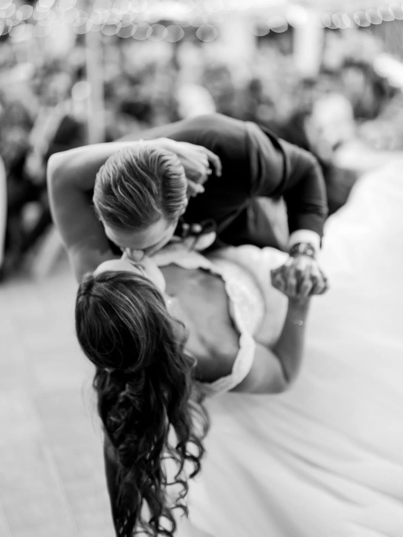 A black-and-white photo of a couple dancing closely; the man leans in to kiss the woman, who has long curly hair and is wearing a wedding dress.