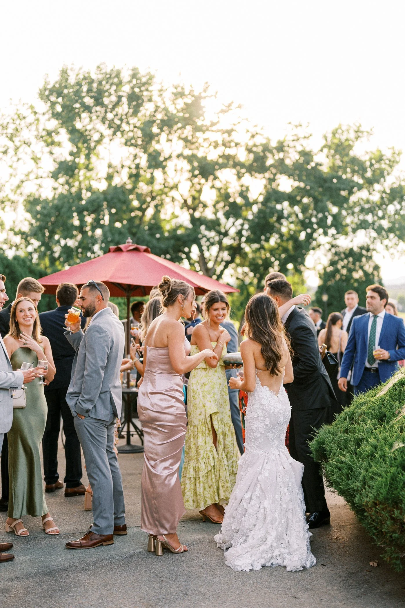 A group of people dressed in formal attire at an outdoor event, possibly a wedding reception, with trees and a red umbrella in the background during sunset.