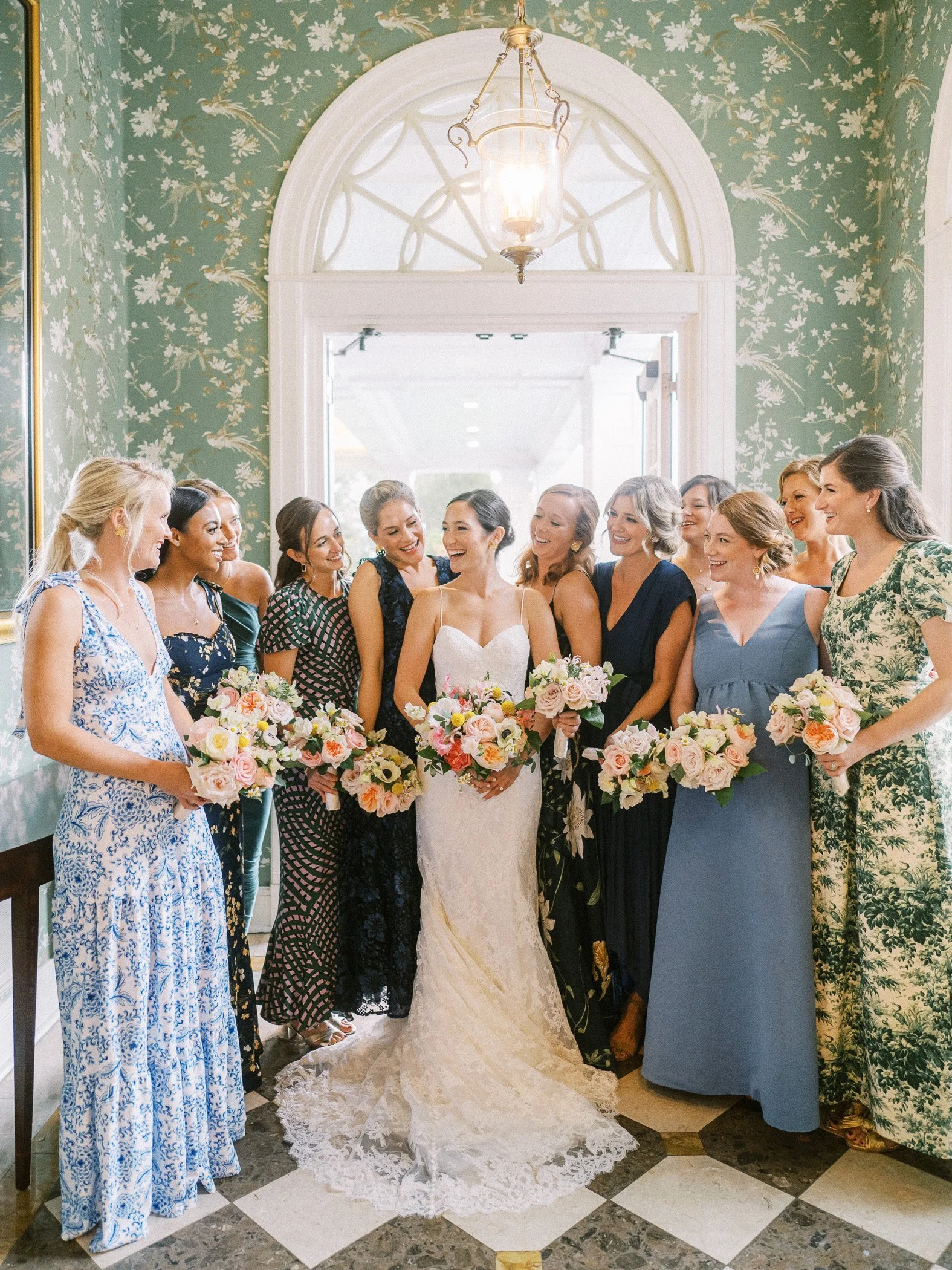 A bride in a white lace wedding dress holding a bouquet, surrounded by smiling women in colorful dresses, each holding bouquets, inside a room with green floral wallpaper and a window with a decorative arch.