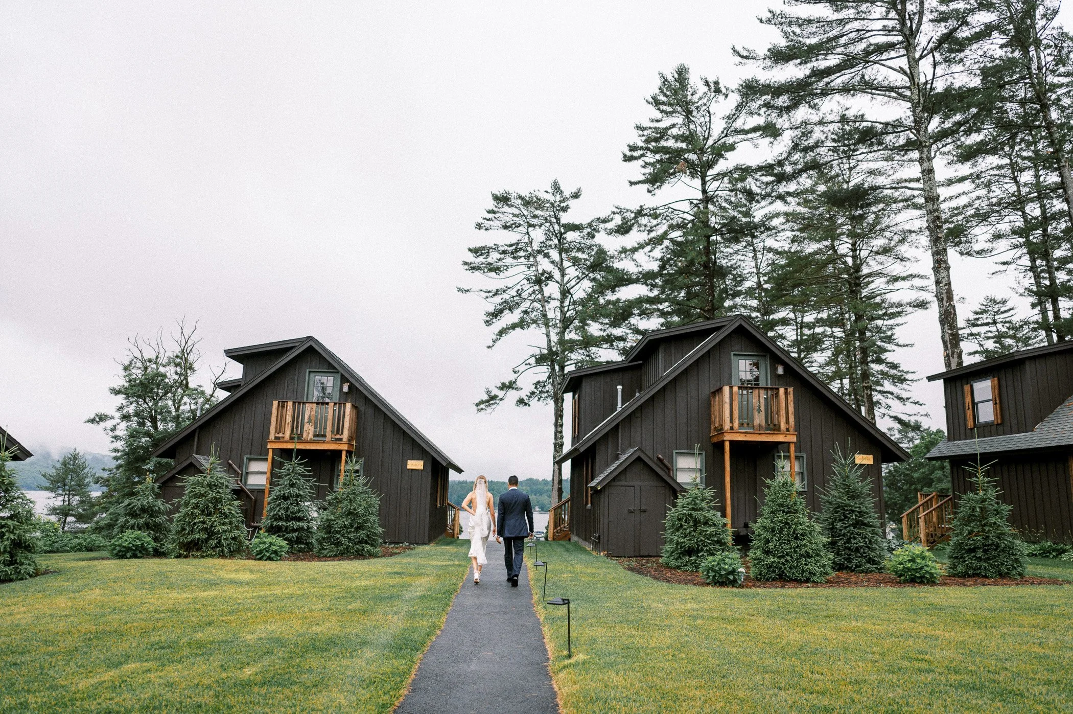 A couple walking down a paved pathway toward two black wooden houses with small balconies, surrounded by green lawn and evergreen trees, in an outdoor setting during daytime.