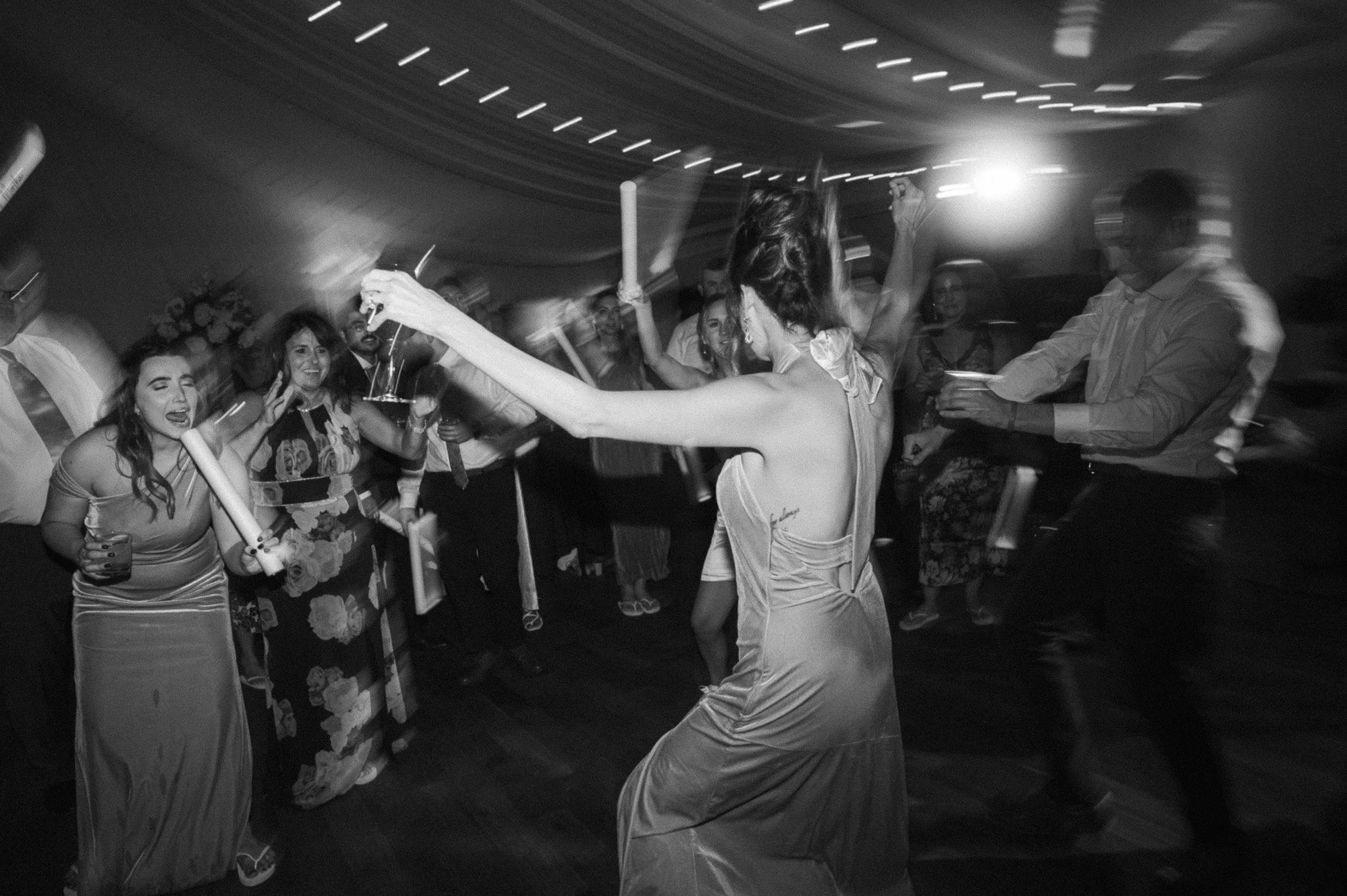 People dancing and celebrating at a party, including a woman in a sleeveless dress in the foreground, some holding drinks, with motion blur indicating lively movement, in a dimly lit venue.