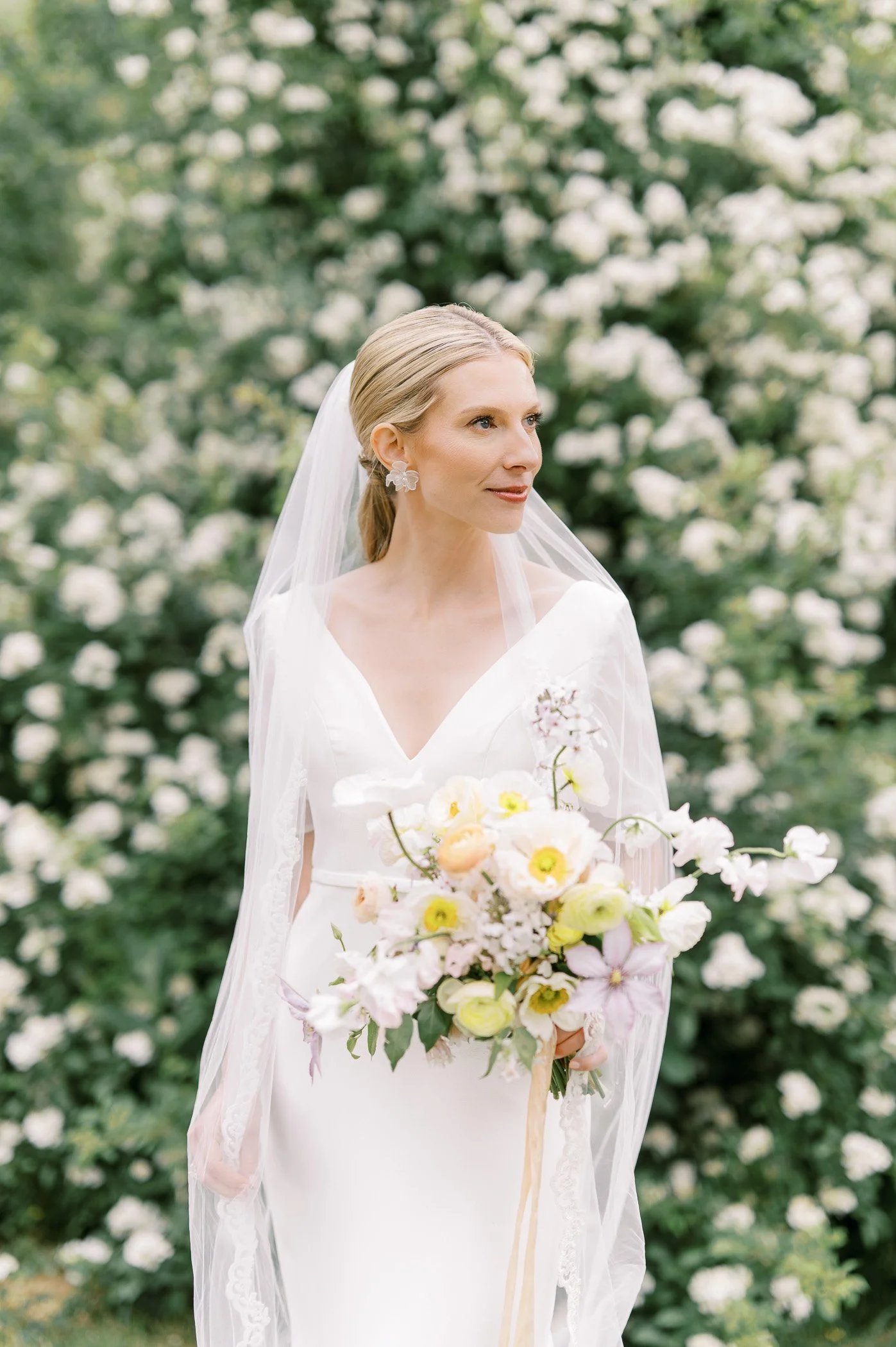 A woman in a white wedding dress holding a bouquet of flowers standing outdoors with a background of blooming white flowers and green foliage.