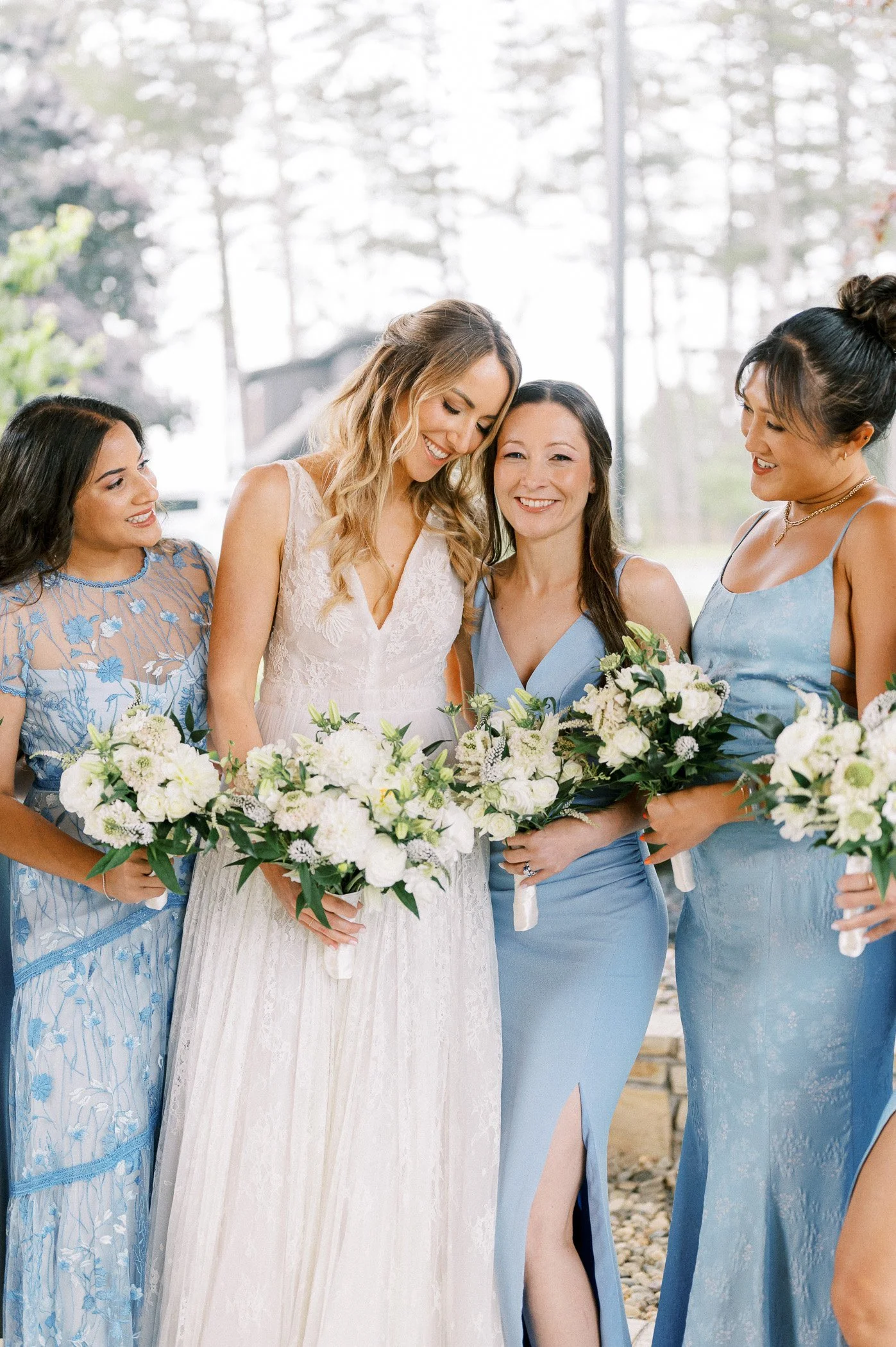 A bride in a white lace dress with blonde hair smiling with four bridesmaids in blue dresses holding bouquets of white flowers.
