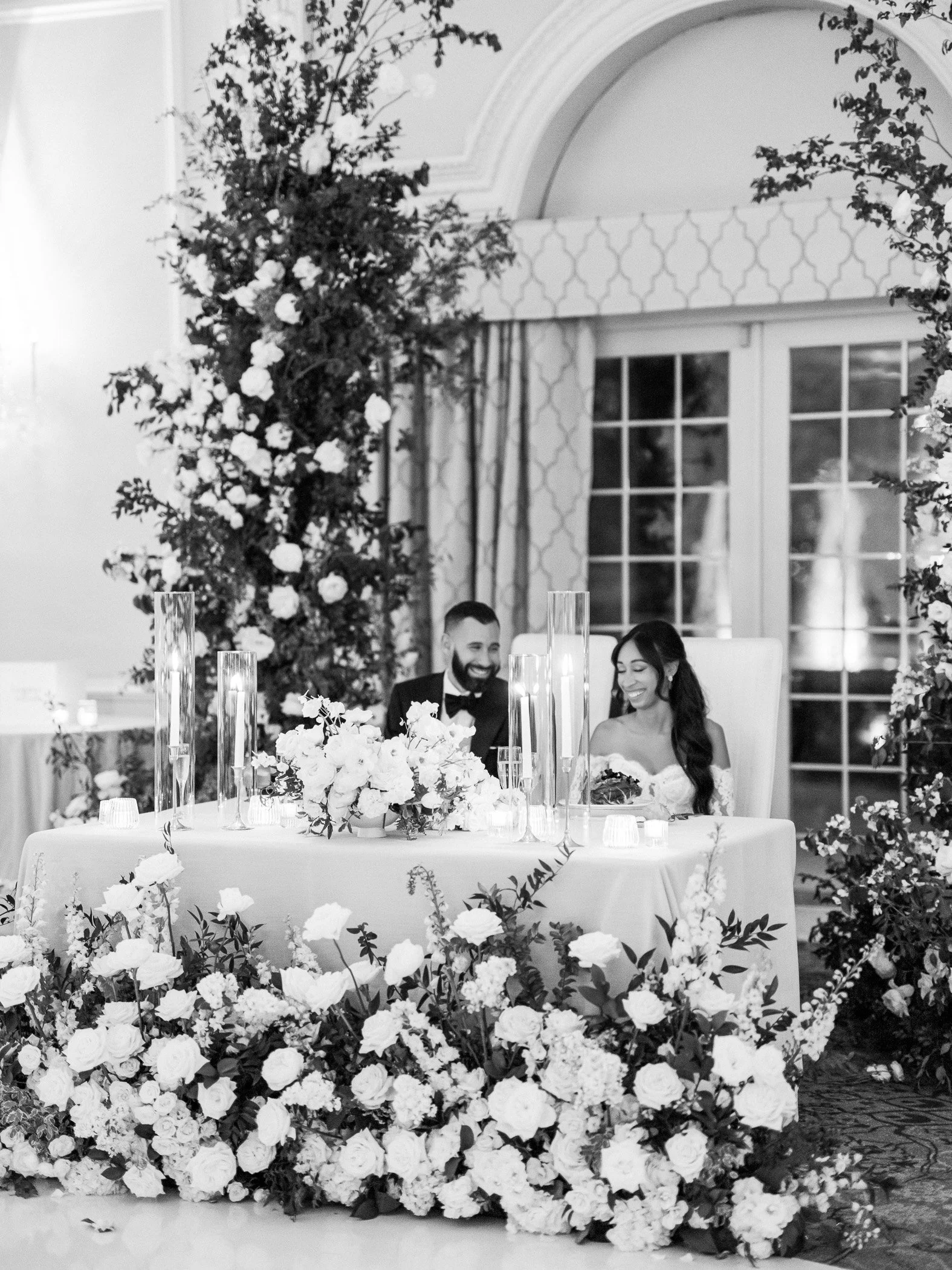 Black and white photo of a wedding reception with a newlywed couple sitting at a decorated head table surrounded by floral arrangements and tall candles.