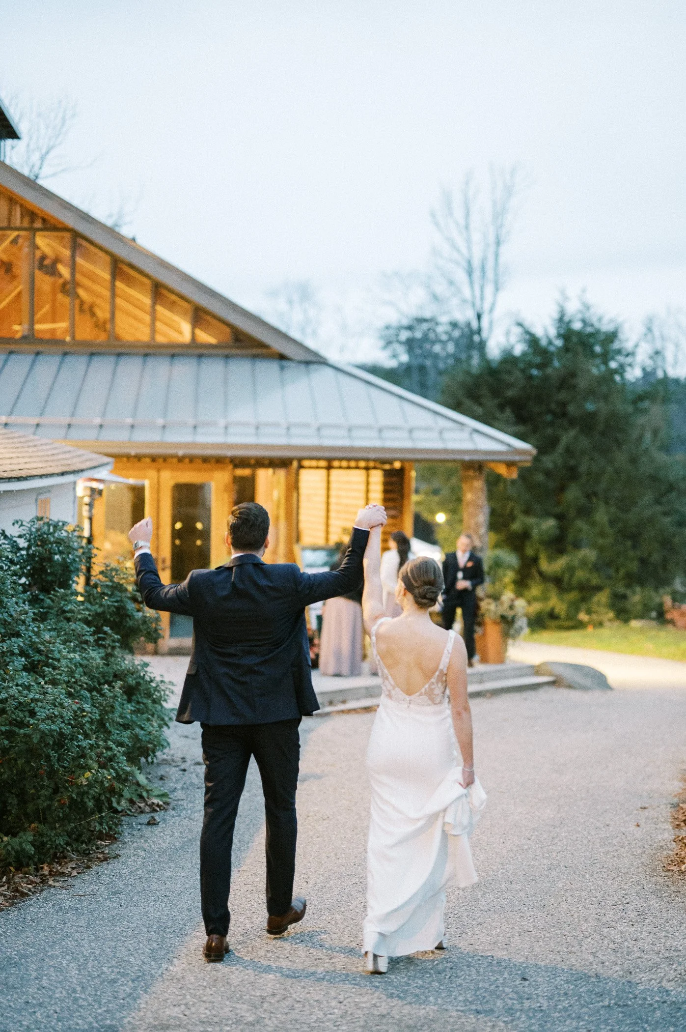 A newlywed couple, wearing formal wedding attire, is walking away from a building, holding hands and celebrating. The groom, in a black suit, is raising one arm while the bride, in a white gown, holds her dress with her other hand. In the background,