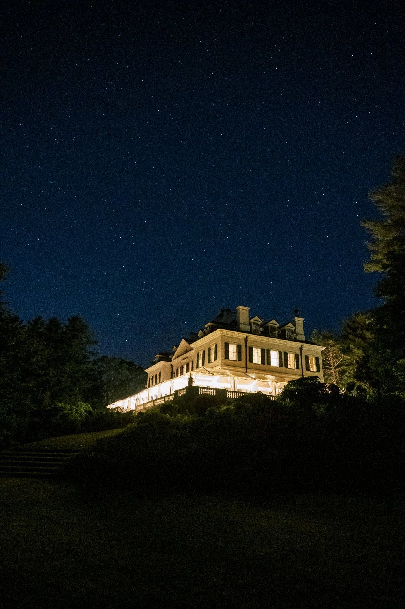 Nighttime view of a large, well-lit house on a hill under a starry sky, surrounded by trees.
