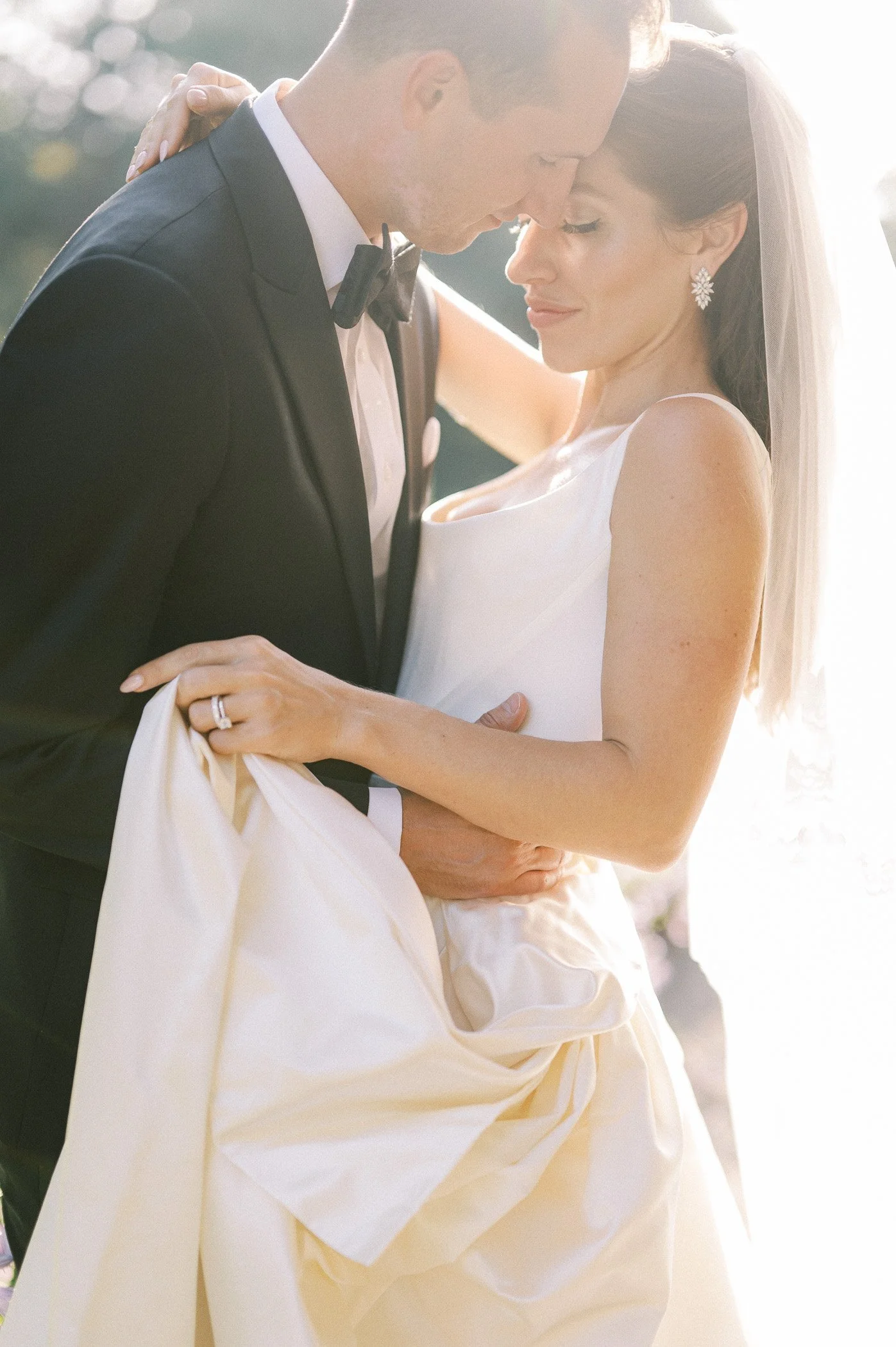 A bride and groom embrace during their wedding, with their foreheads touching and eyes closed, outdoors in bright sunlight.