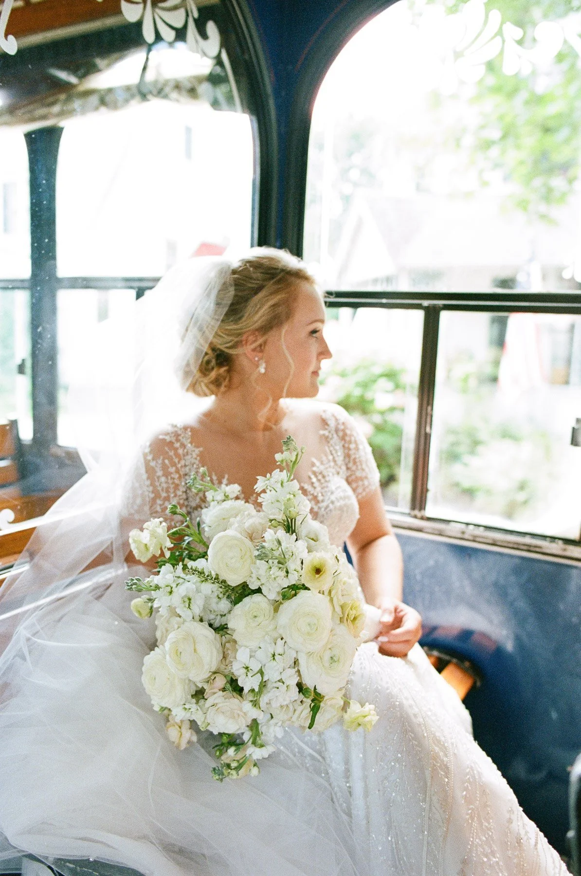 A bride in a white wedding dress with lace details, holding a large bouquet of white flowers, sitting in a vintage vehicle with open windows, looking out.