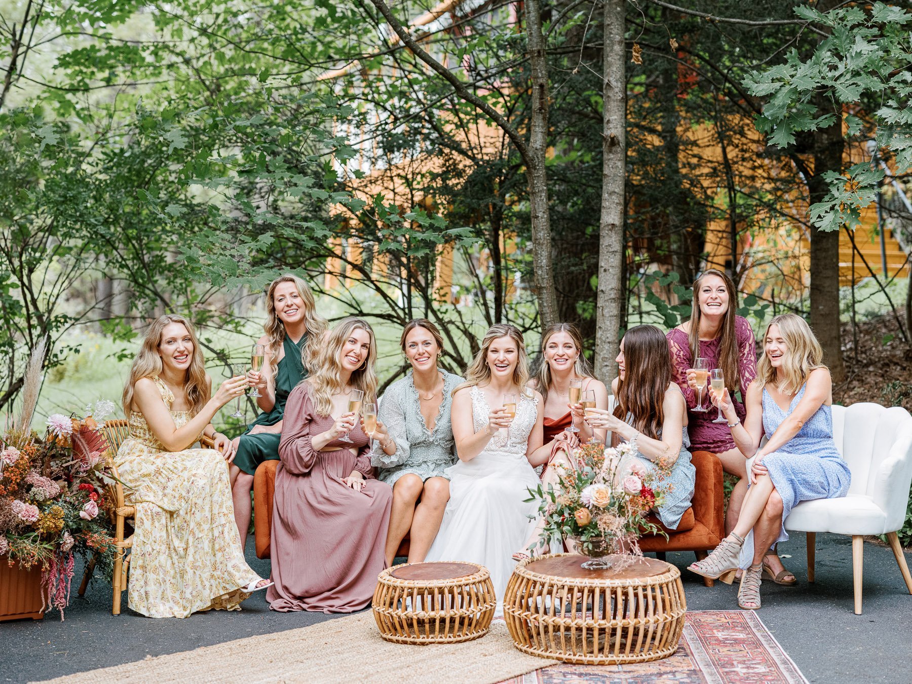 Group of women in elegant dresses sitting outdoors on a sofa and chairs, holding glasses of champagne, celebrating a special occasion in a wooded area with trees and greenery.