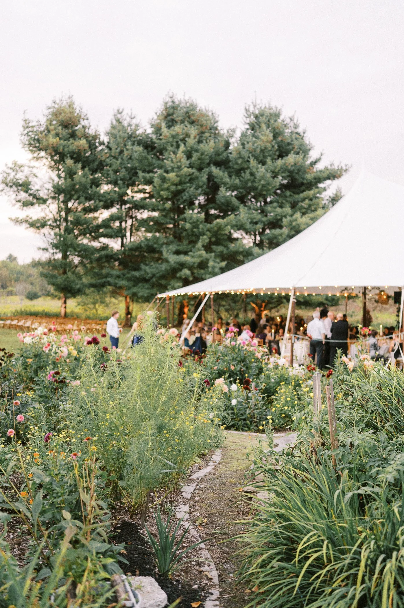 Outdoor wedding reception tent surrounded by flower garden with guests and staff inside.