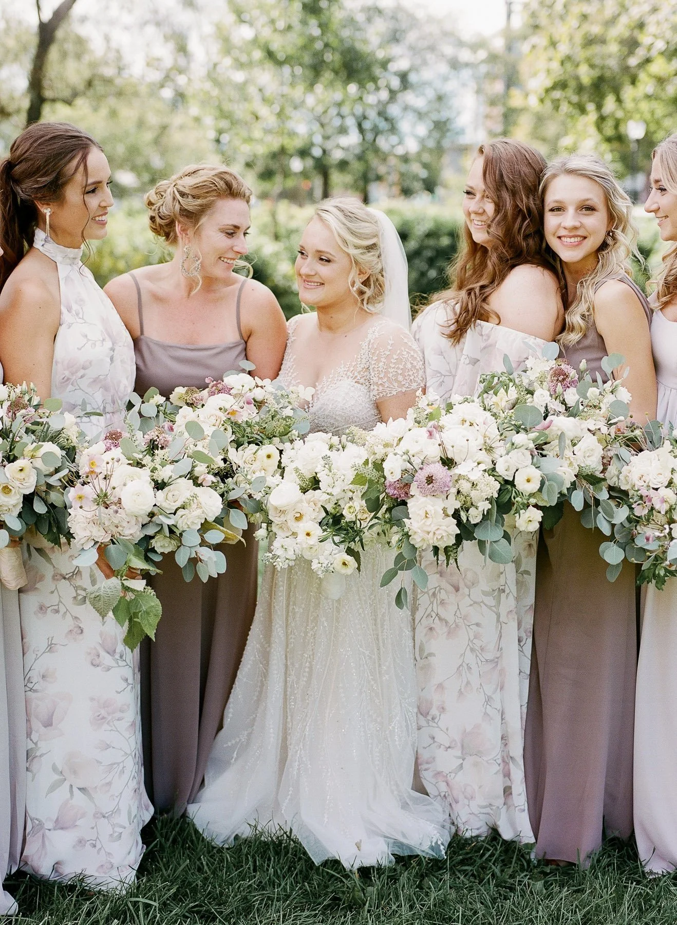 A bride and six bridesmaids standing outdoors with large floral arrangements, smiling and looking at each other.